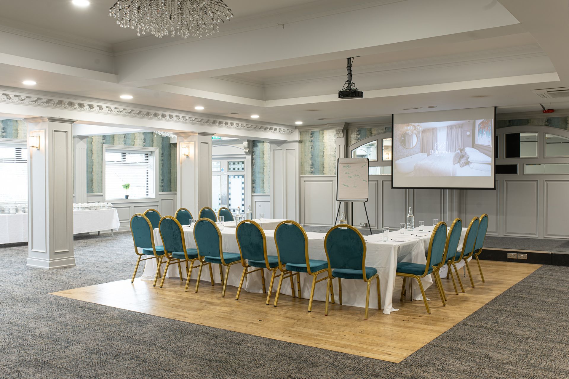 Elegant banquet hall with a long white-draped table, teal chairs, and a projector screen at the front.