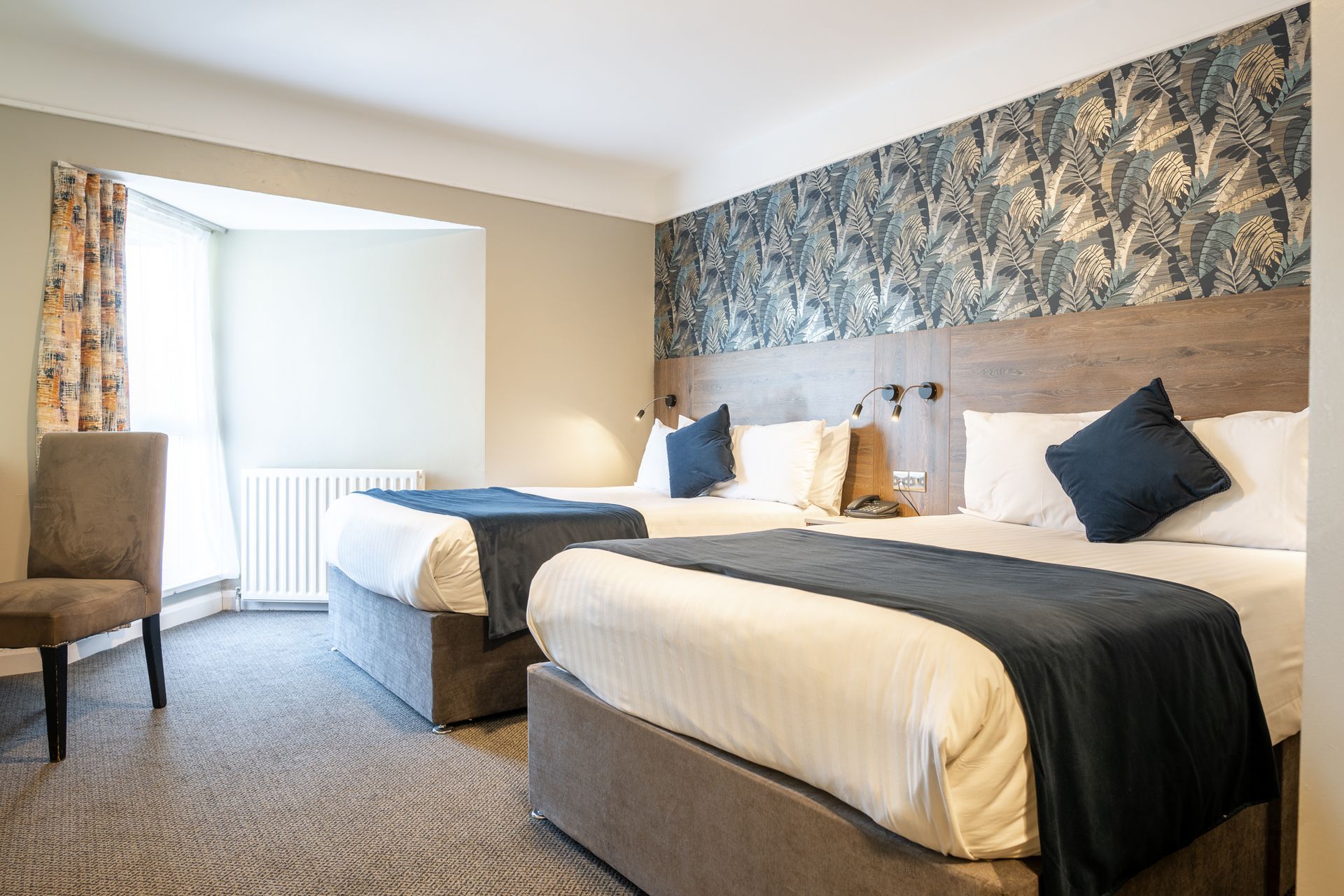 Hotel room with two beds, patterned wallpaper, and a window with curtains and a chair.