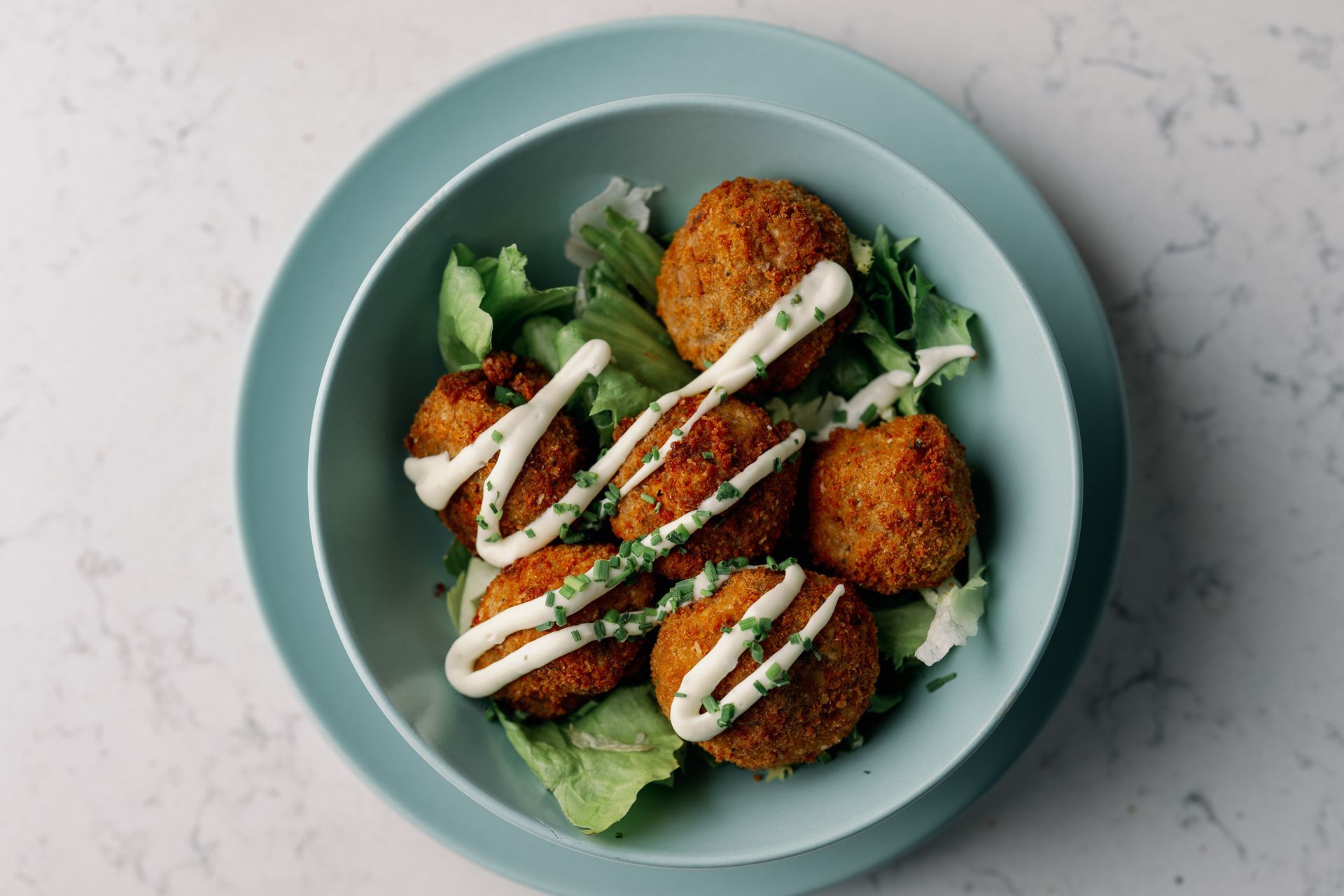 Top-down view of crispy falafel on lettuce in a teal bowl, drizzled with white sauce