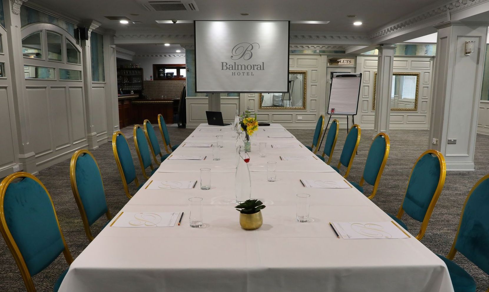 Conference room with a long white table, teal chairs, and small potted plants down the center.