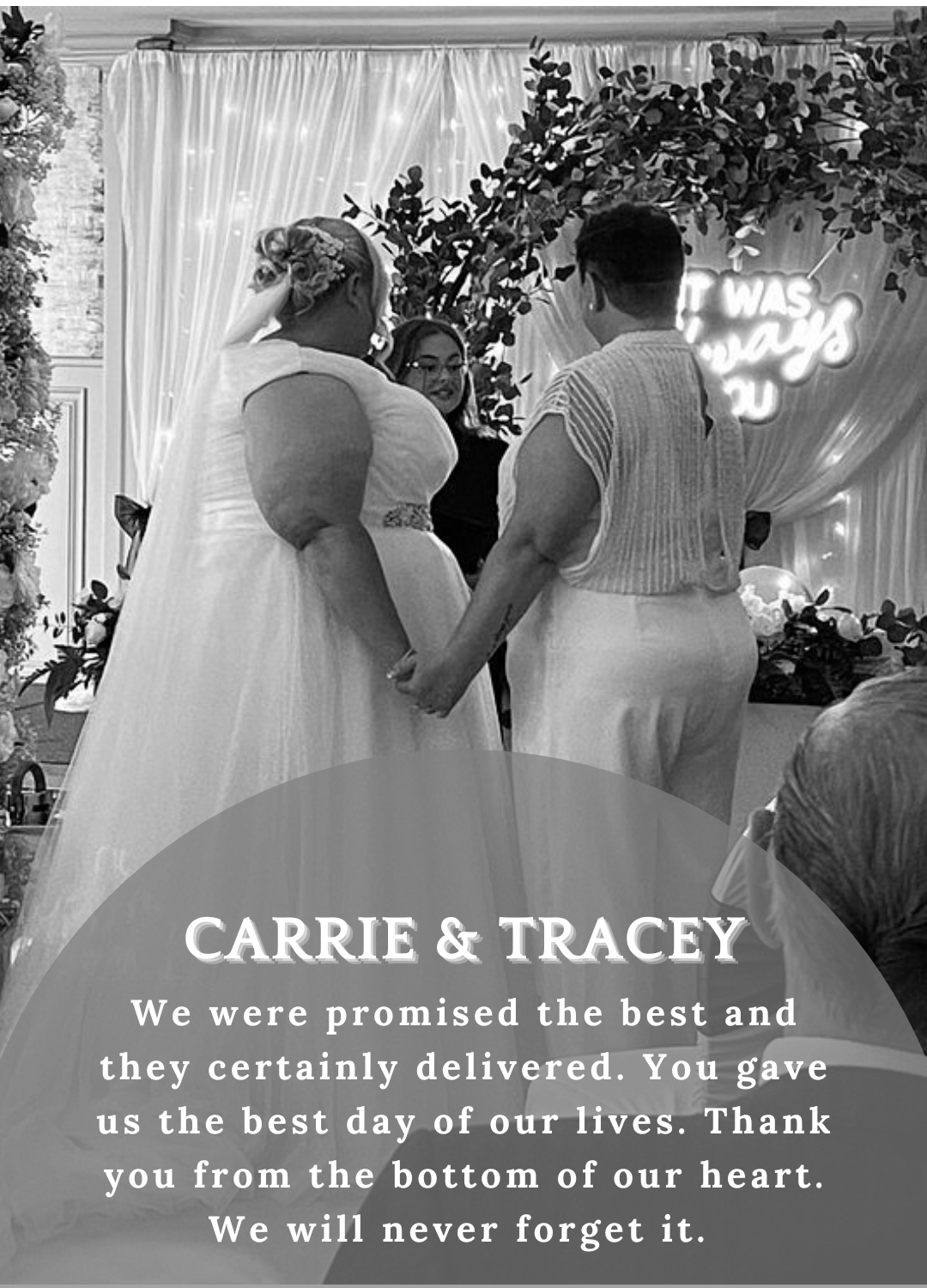 Two people in white wedding attire holding hands at a decorated reception backdrop with a tribute sign.