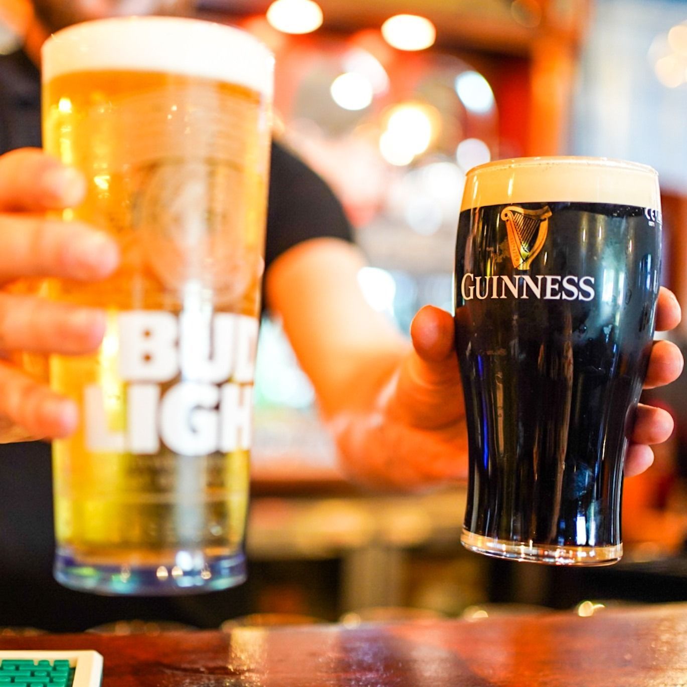 Hands holding Guinness and Bud Light pints in a warm, bar-like setting