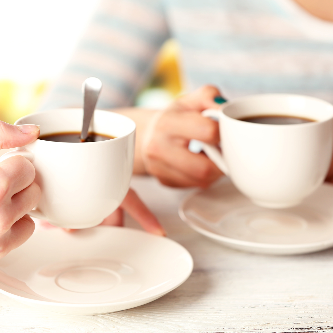 Two people holding white coffee cups at a bright table, with spoons and saucers visible