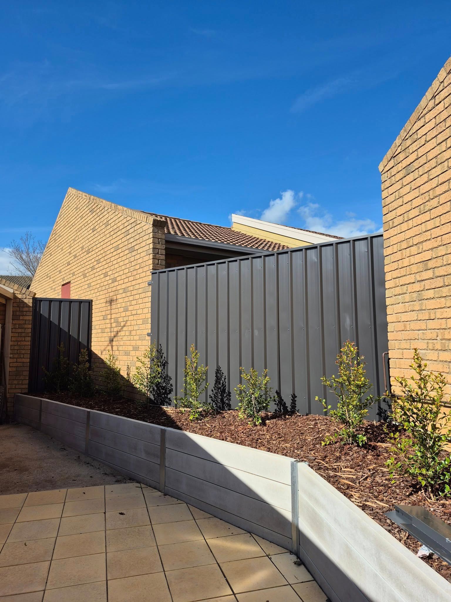 Brick buildings and gray fence next to a garden with small shrubs under a bright blue sky — Precision Fencing & Landscaping in Thurgoona, NSW