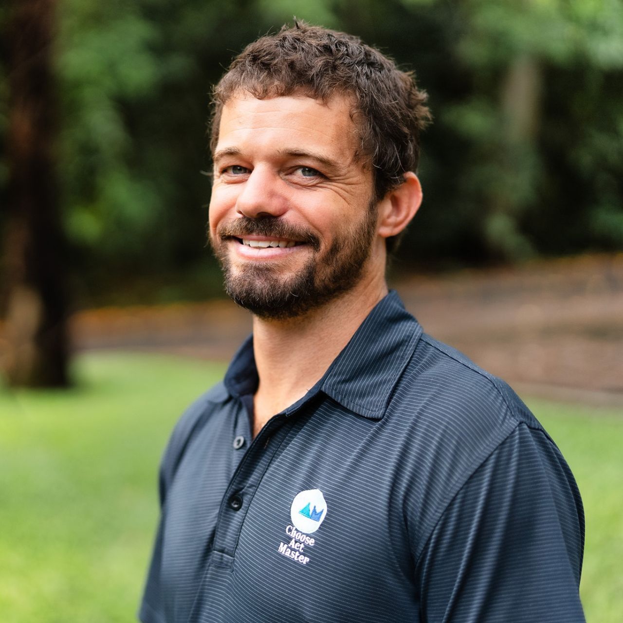 Cameron Matthew from Choose Act Master Performance Coaching - a man with a beard is wearing a blue shirt and smiling .