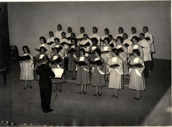Coral em trajes claros, com partituras nas mãos, regido no palco na primeira récita oficial, em 1961