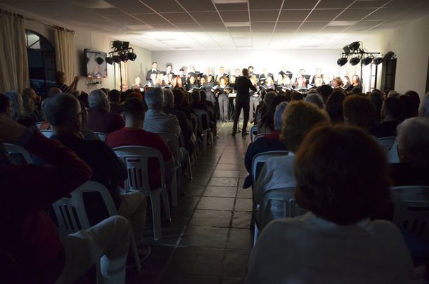 Público sentado no auditório da sede, de costas, assistindo ao coral ao fundo sob refletores