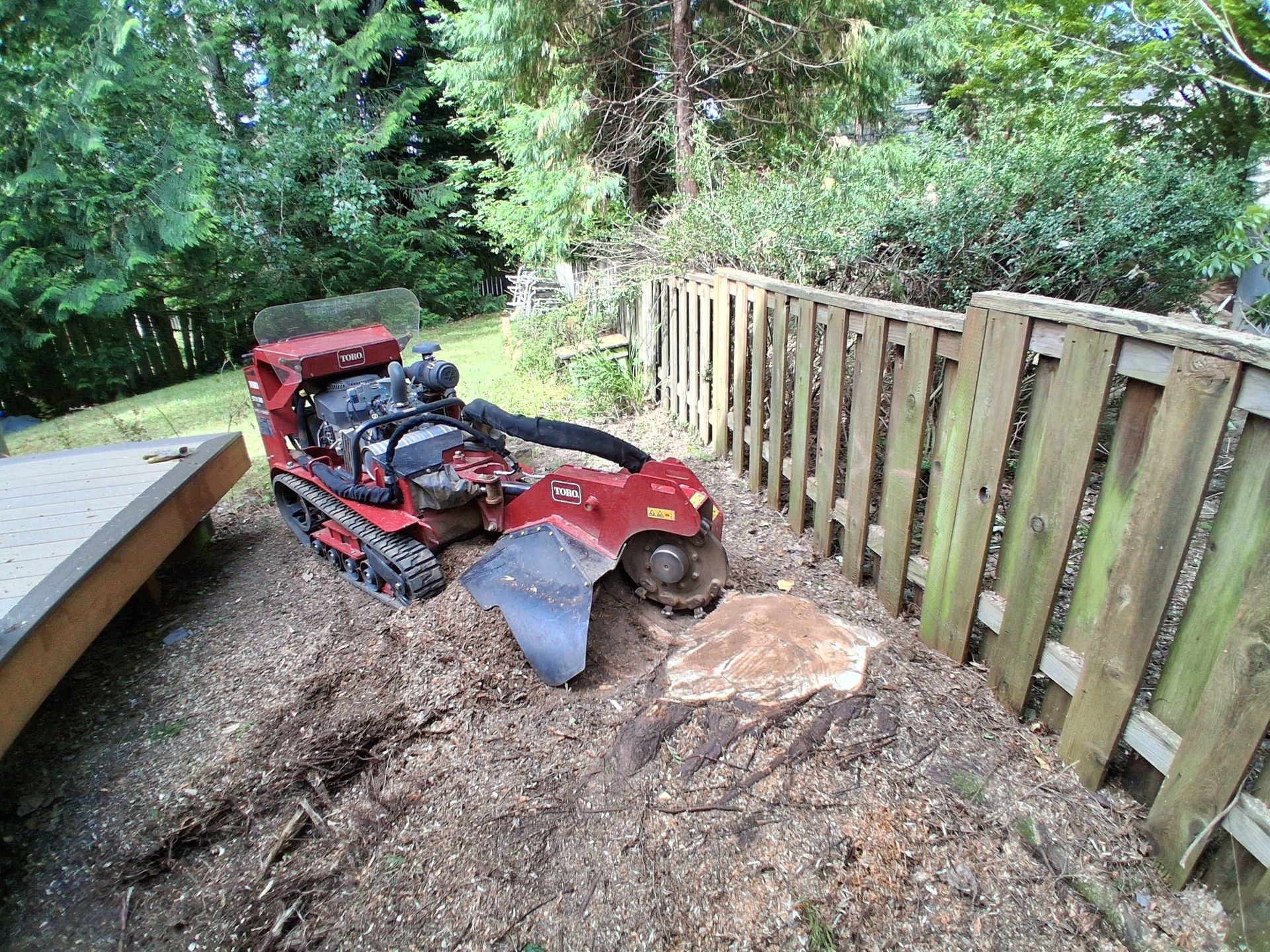Red lawn mower stuck in muddy trench beside a wooden fence and trees