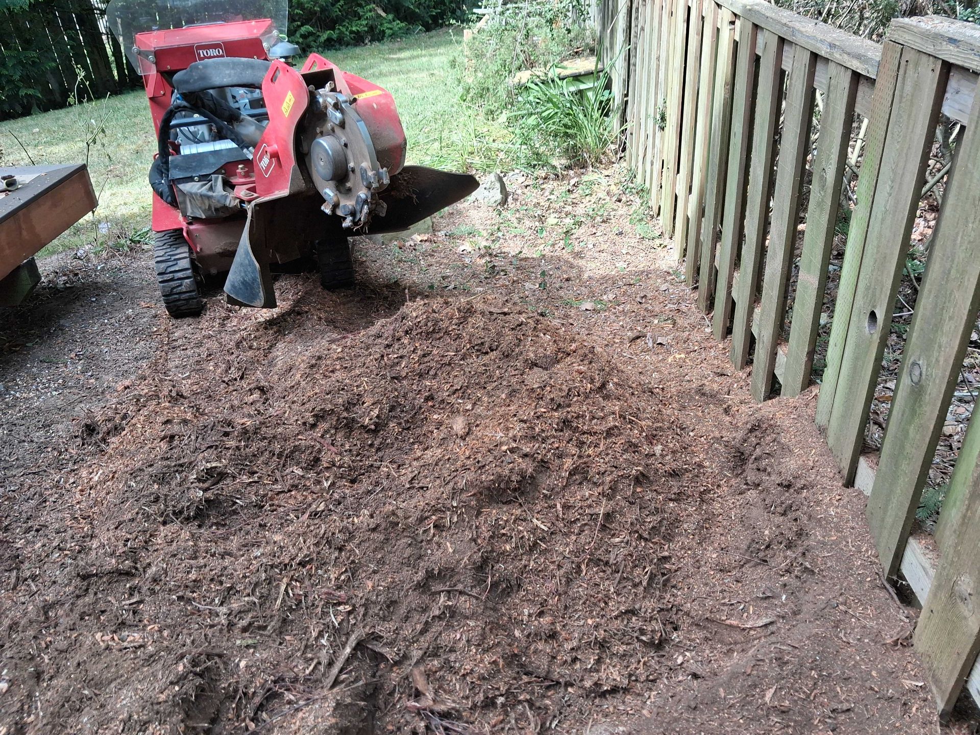 Red tractor spreading mulch beside a wooden fence in a garden path area.