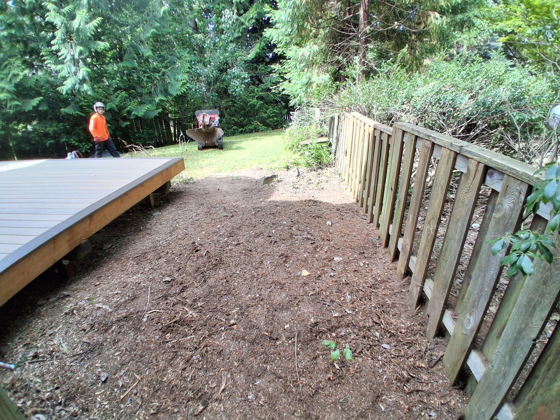 Wooded backyard path with mulch, a wooden deck on the left, fence on the right, and a person in orange in the distance