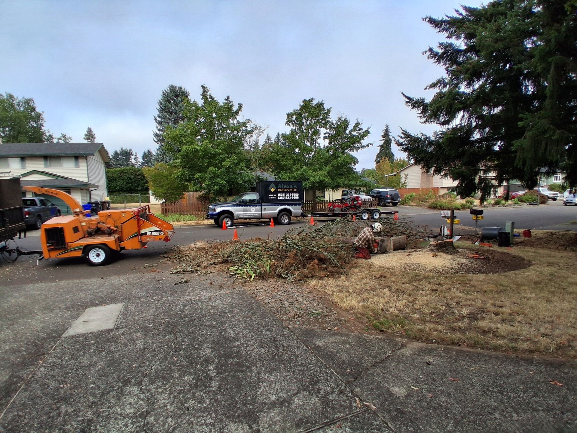 Orange wood chipper and debris pile in a driveway near parked vehicles and trees.