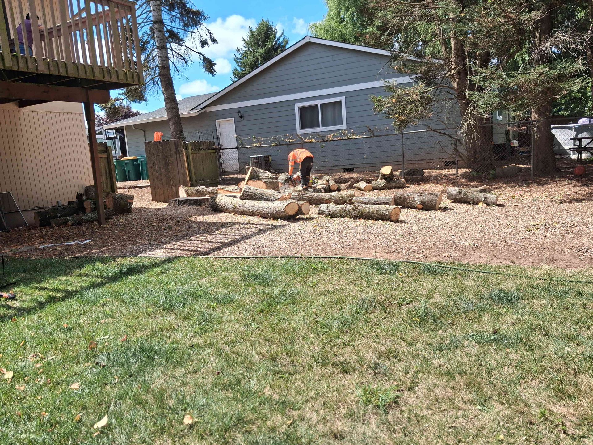Backyard with green lawn, rock garden, and gray house under trees; person bending near fire pit