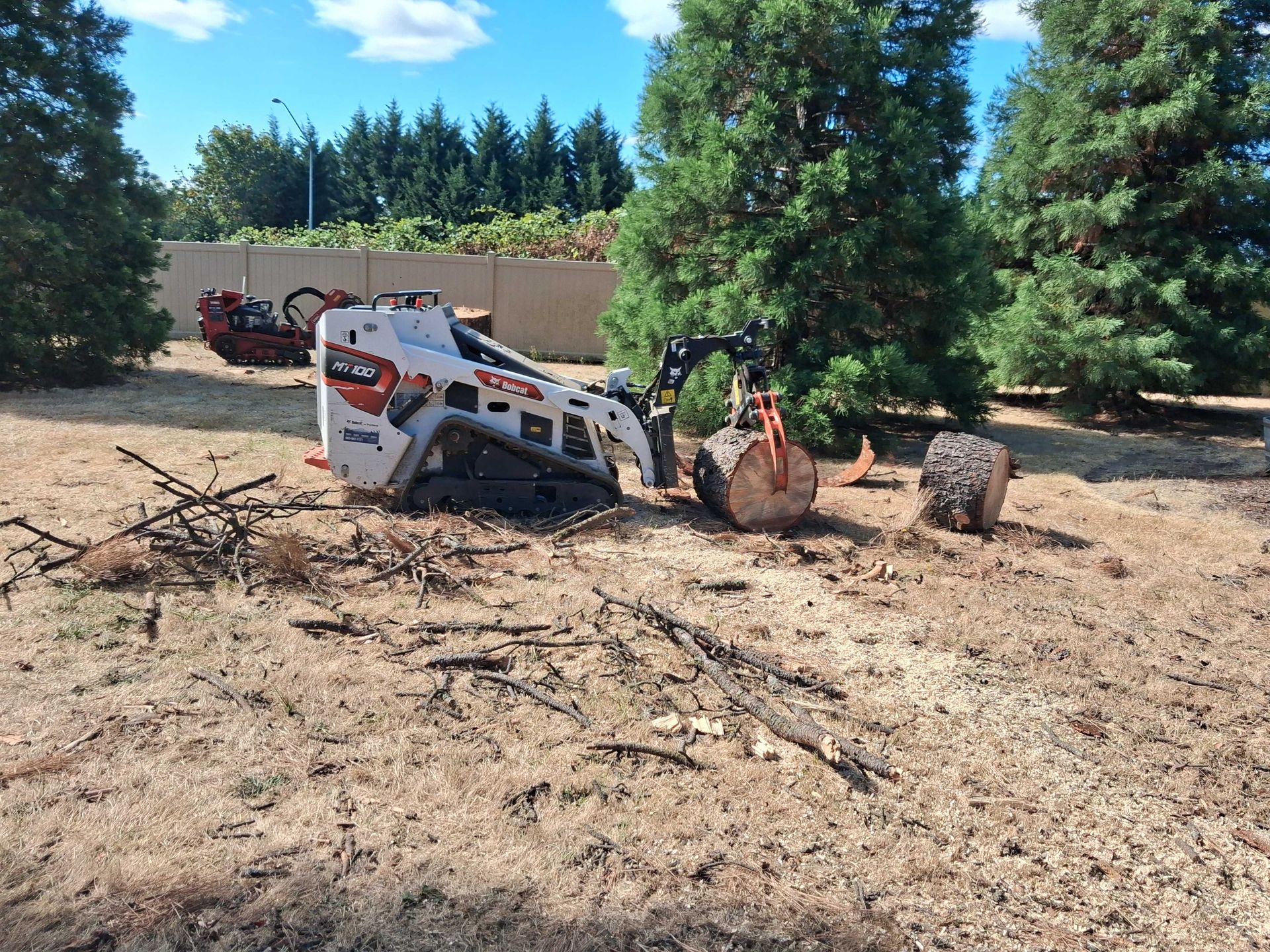 Overturned skid steer in a dirt lot with scattered branches and trees in the background