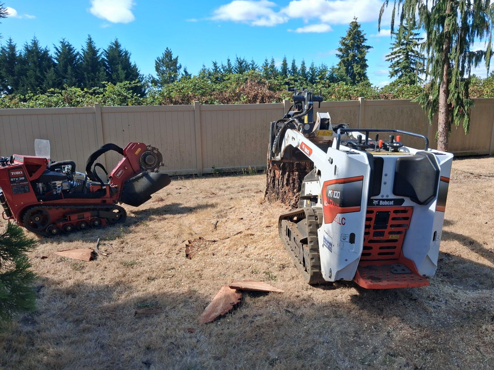 Overturned white skid steer and red machine in a dirt yard beside a fence and trees