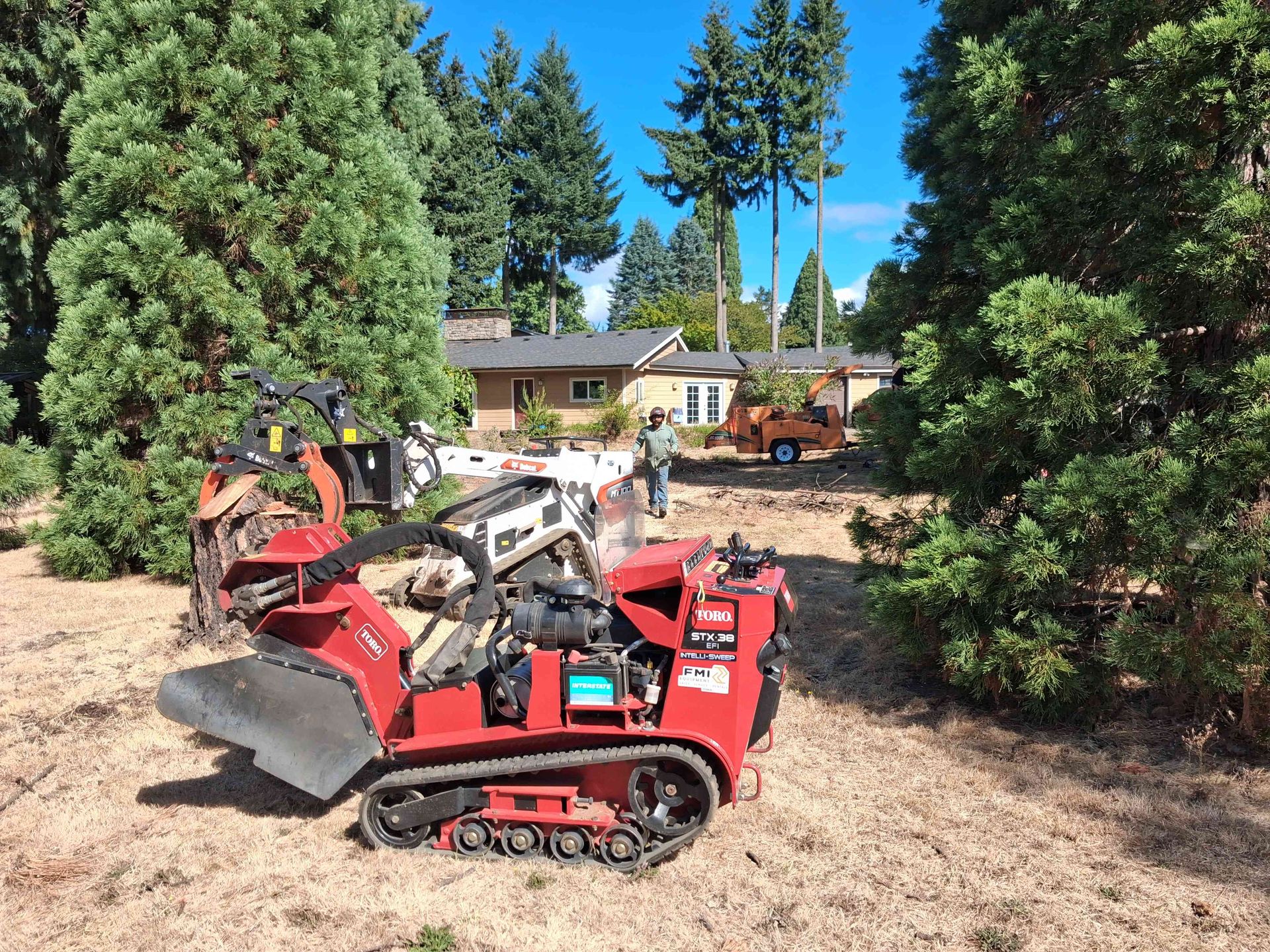 Red tracked wood chipper in a wooded yard near a house and scattered debris