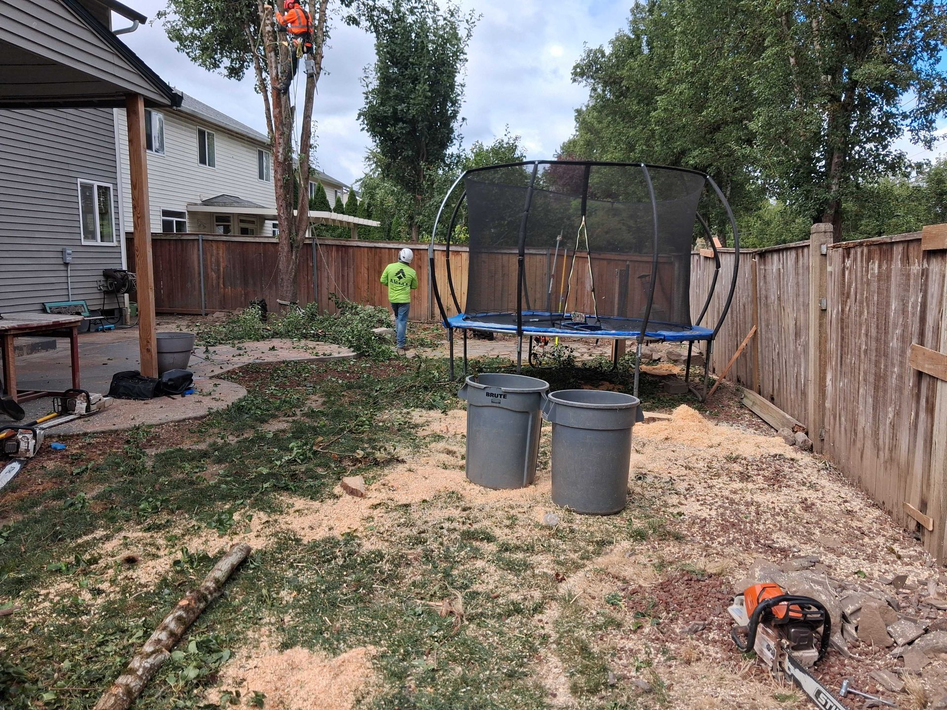 Backyard with dirt ground, trampoline, trash bins, and a worker near the fence.