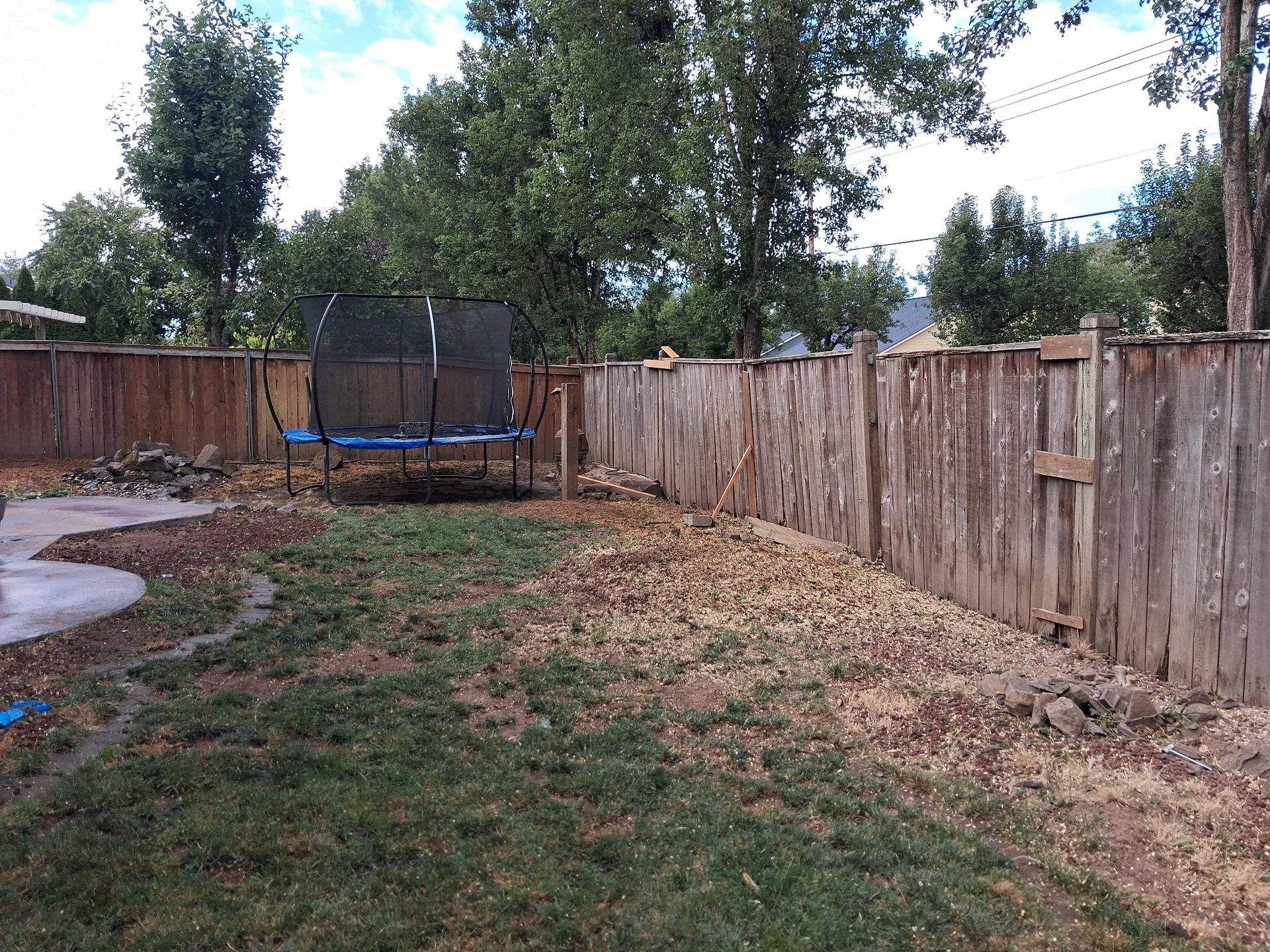 Backyard with a weathered wooden fence, trampoline, trees, and patchy grass under a cloudy sky