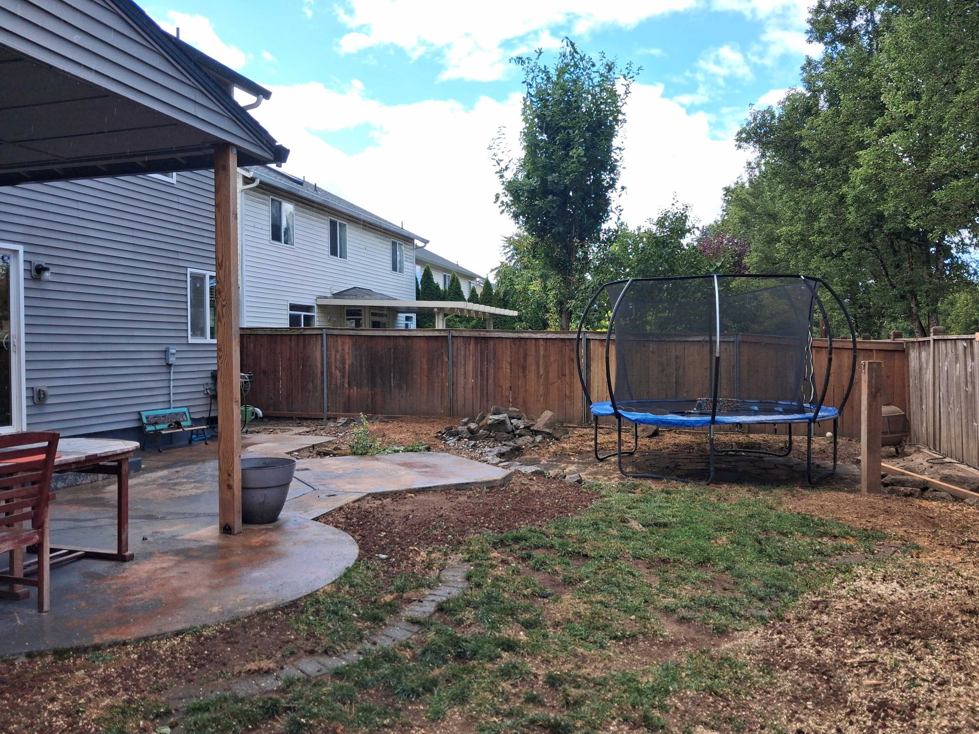 Backyard with trampoline, wooden fence, patio, and a house under a partly cloudy sky