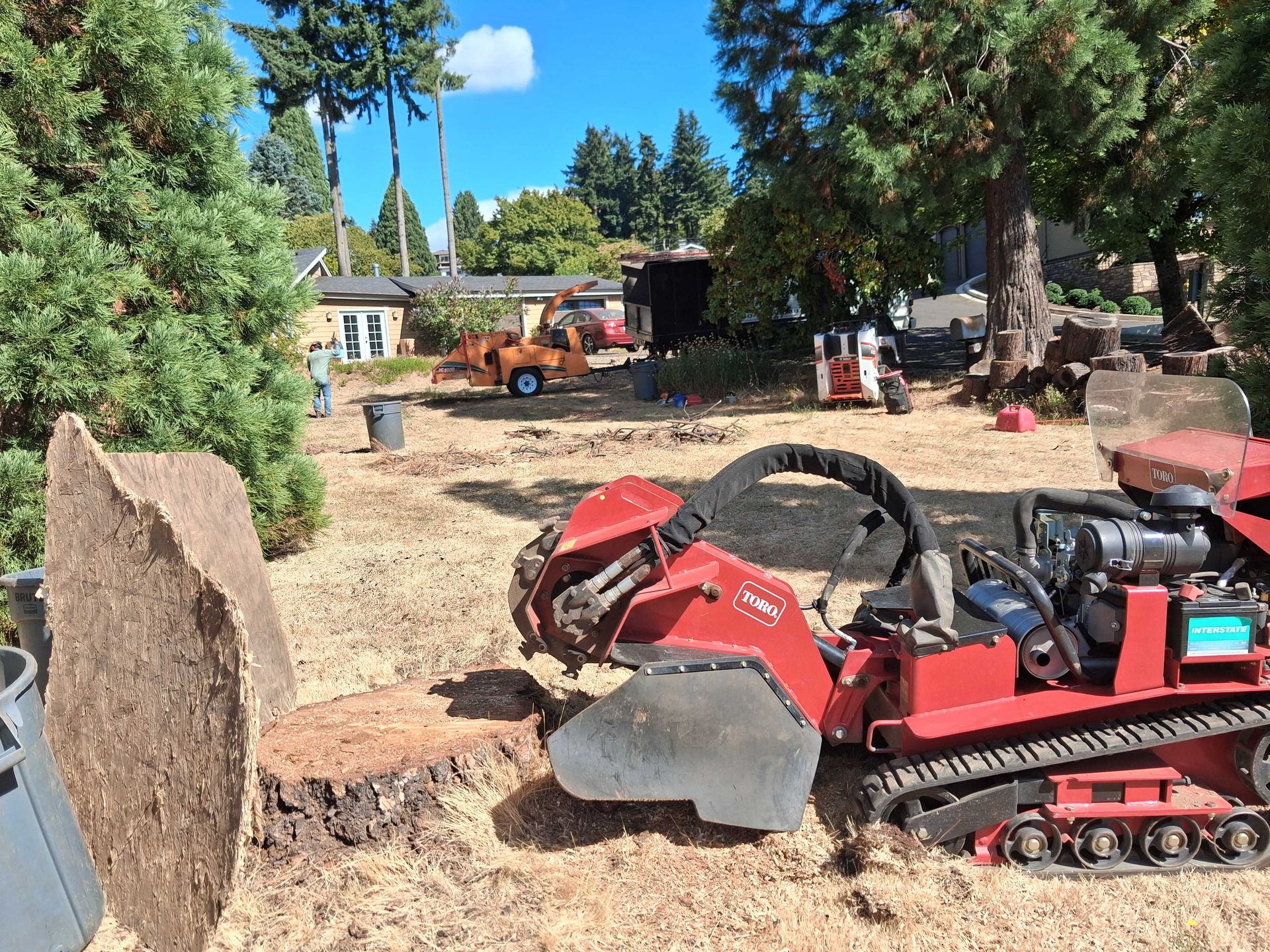 Red compact track loader digging a trench in a yard with trees, dirt piles, and workers in the background
