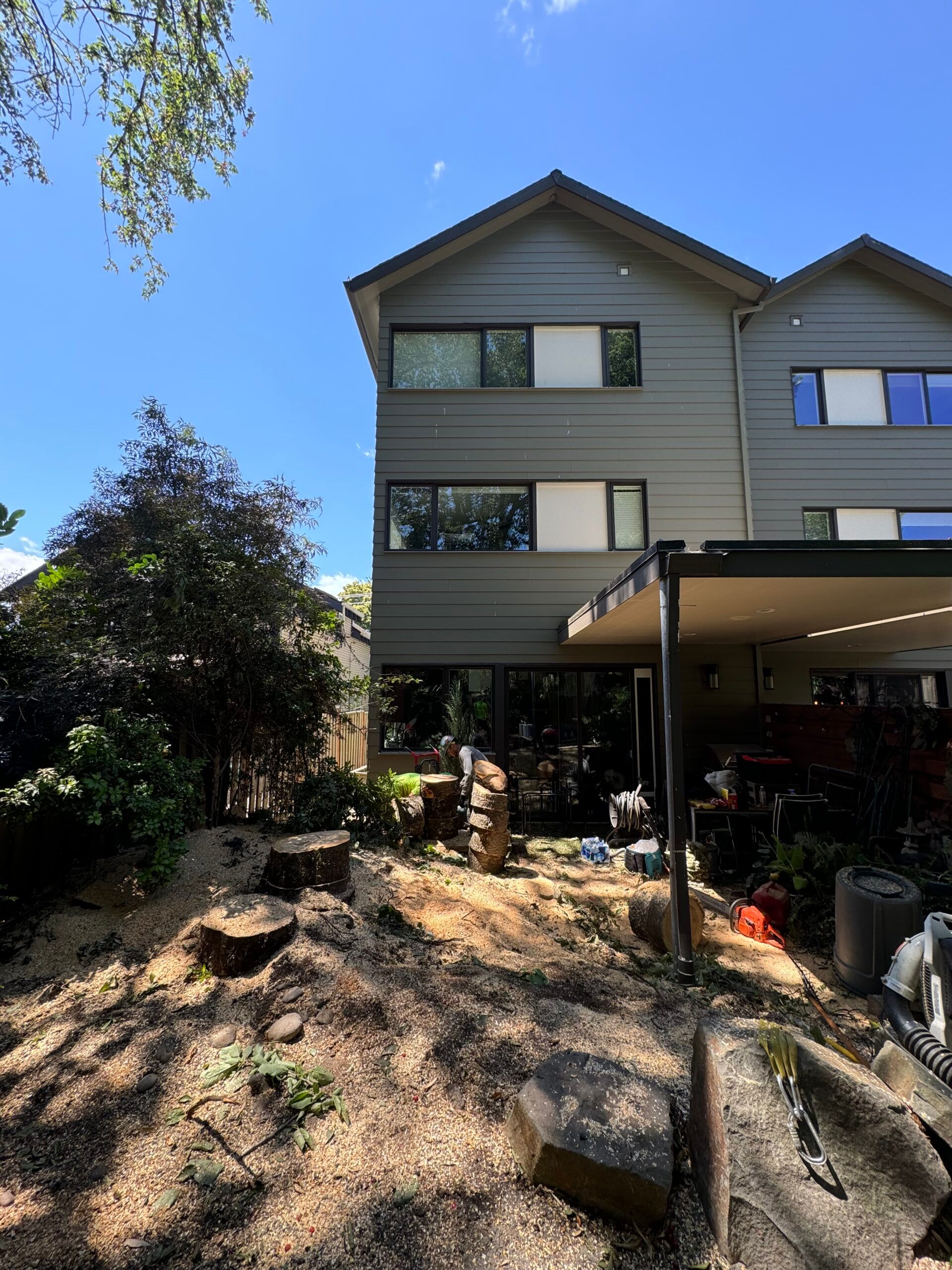 Three-story beige townhouse with covered patio, shaded garden bed, and blue sky overhead