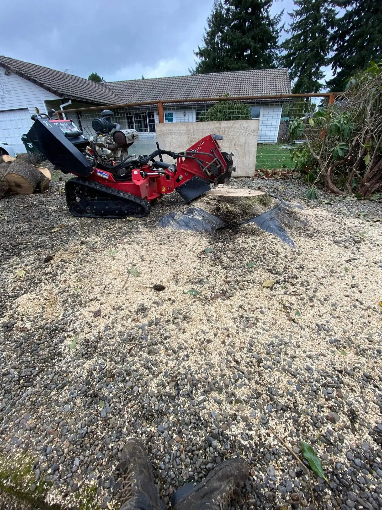 Red excavator stuck in a muddy yard beside a small house and trees.