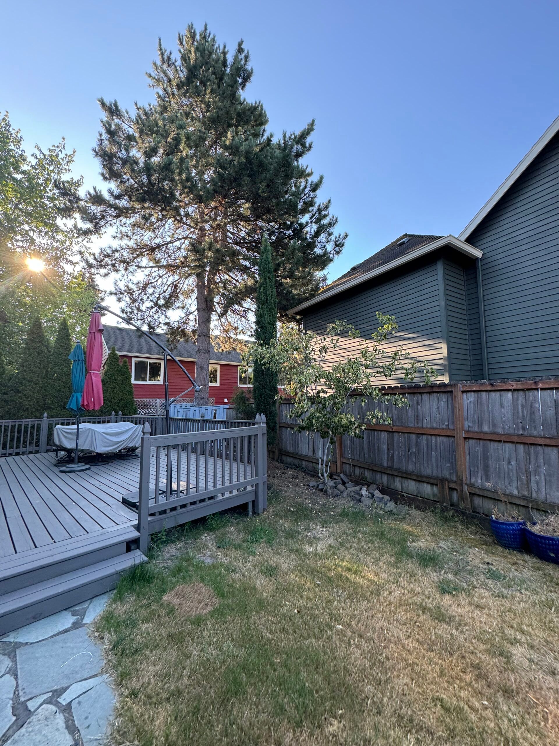 Backyard deck with patio umbrella, fence, and tall tree beside a house at sunset