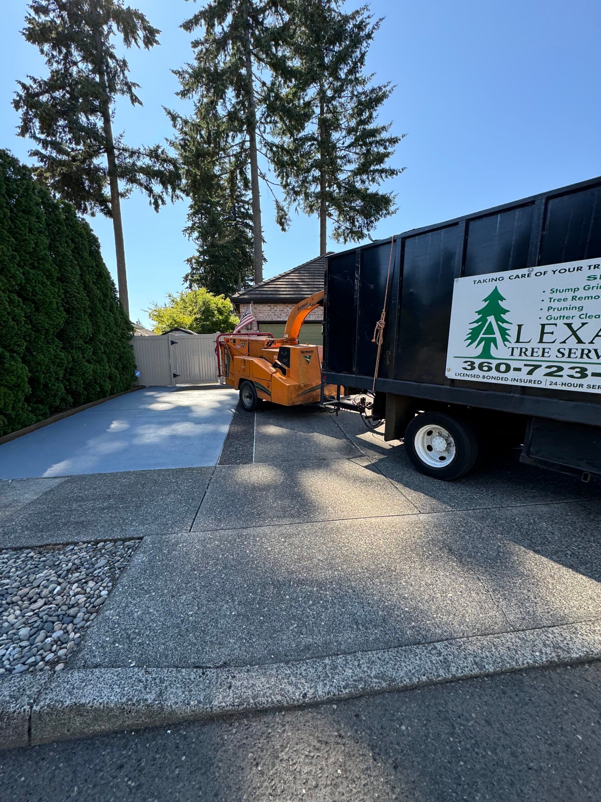 Orange forklift beside black truck in a driveway, with tall trees and a hedge nearby.