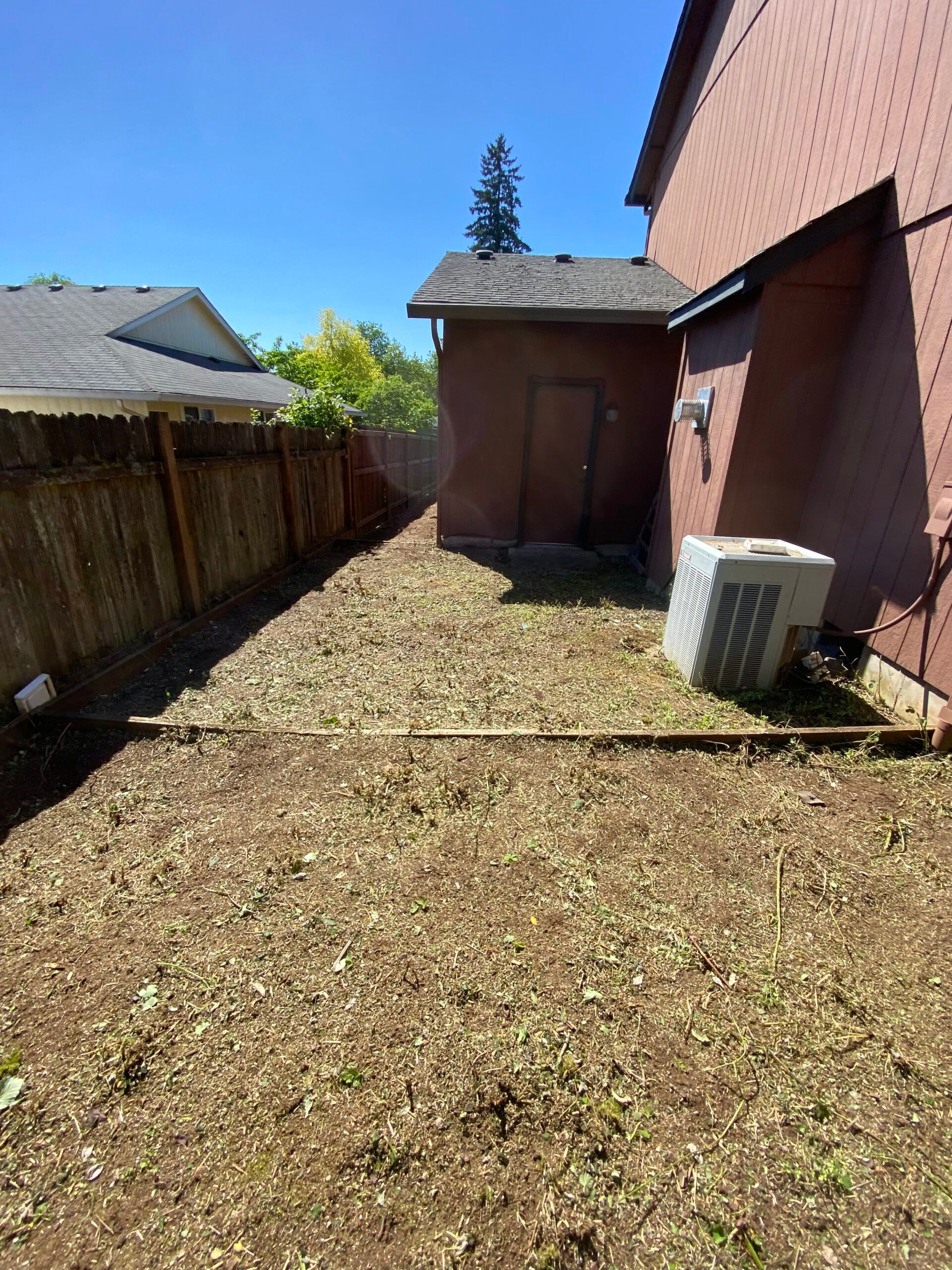 Backyard beside a brick house, with a small shed, wooden fence, and gravel ground under a blue sky.