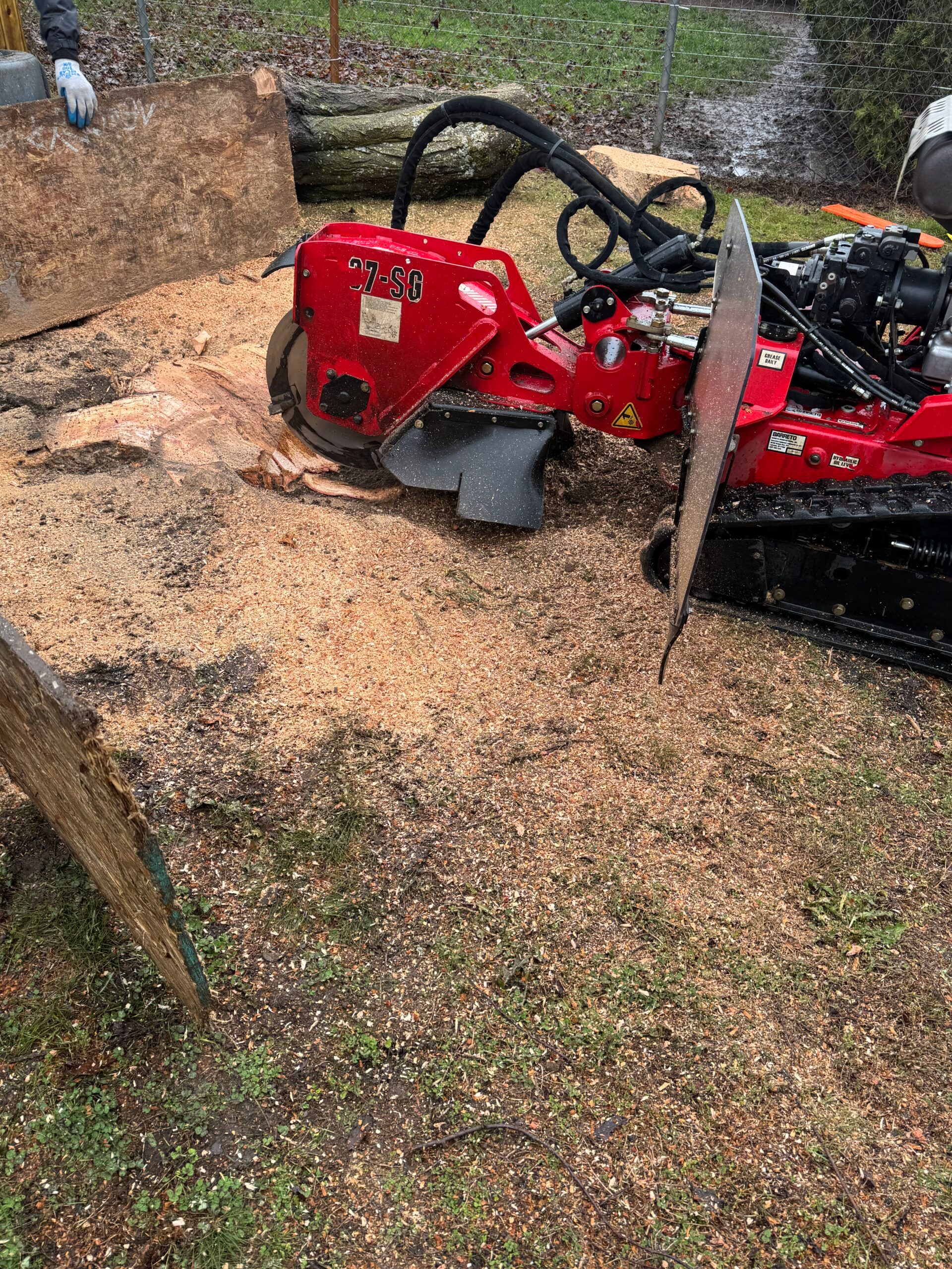 Red off-road vehicle with roll cage parked on a gravel yard beside a wooden fence.