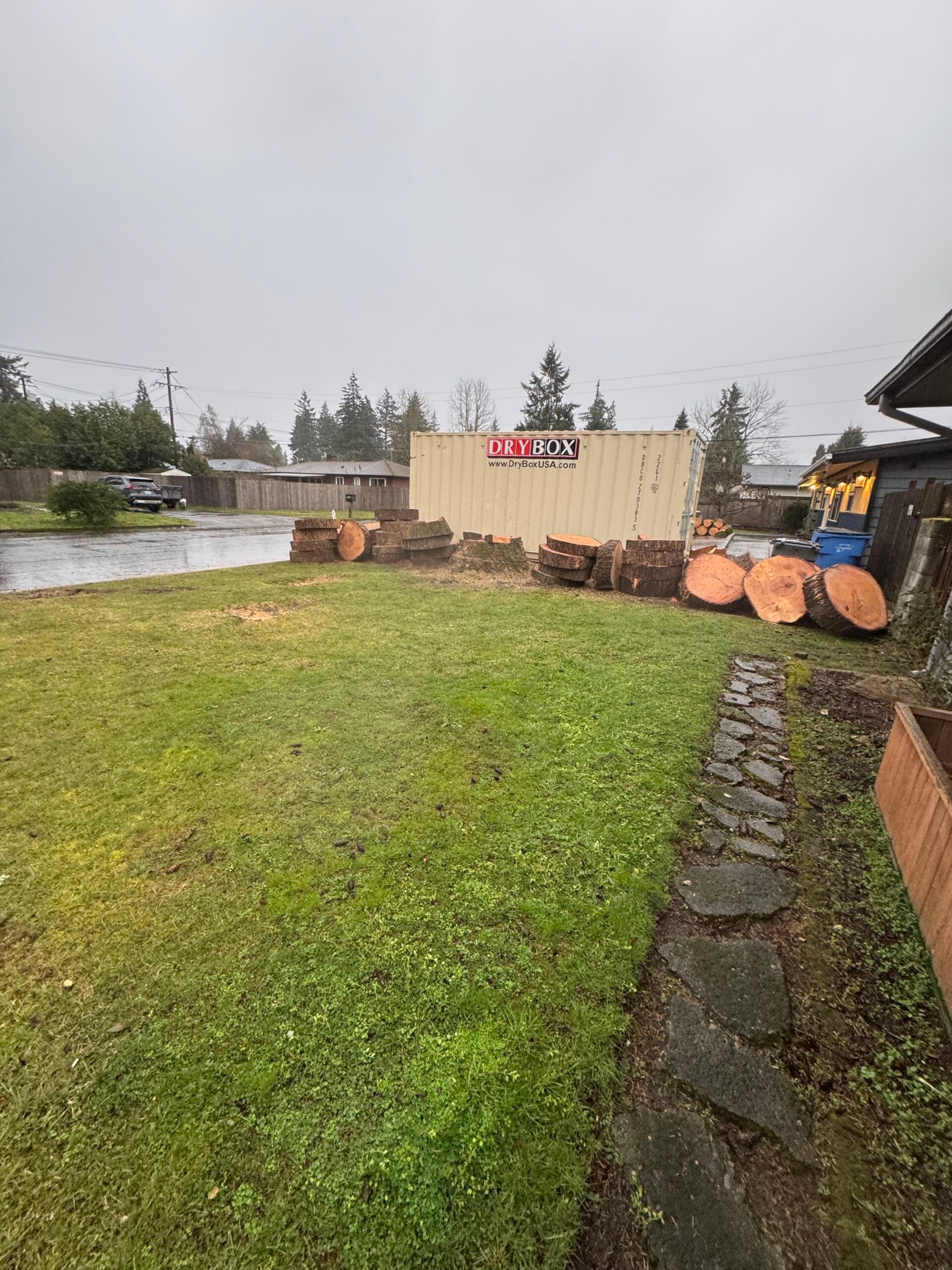 Rainy backyard with green lawn, stone path, woodpile, and shed by the water