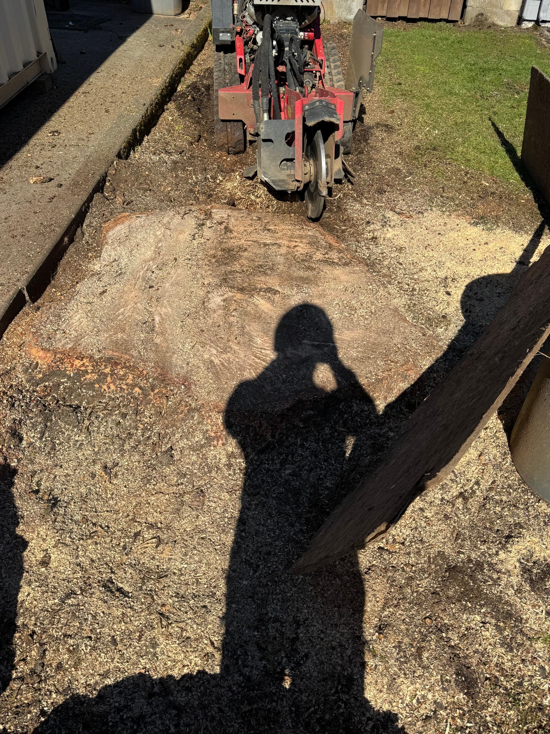 Mini excavator in a gravel yard, digging a trench beside retaining walls, with a person’s shadow in the foreground