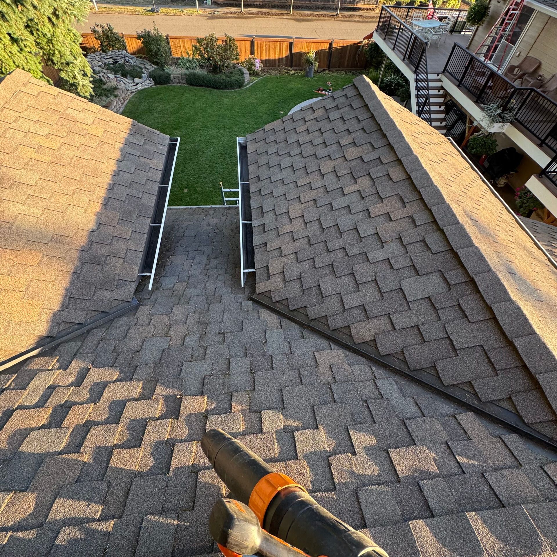 Gabled rooftops framing a green backyard, viewed from a roof ridge with a hand tool in the foreground