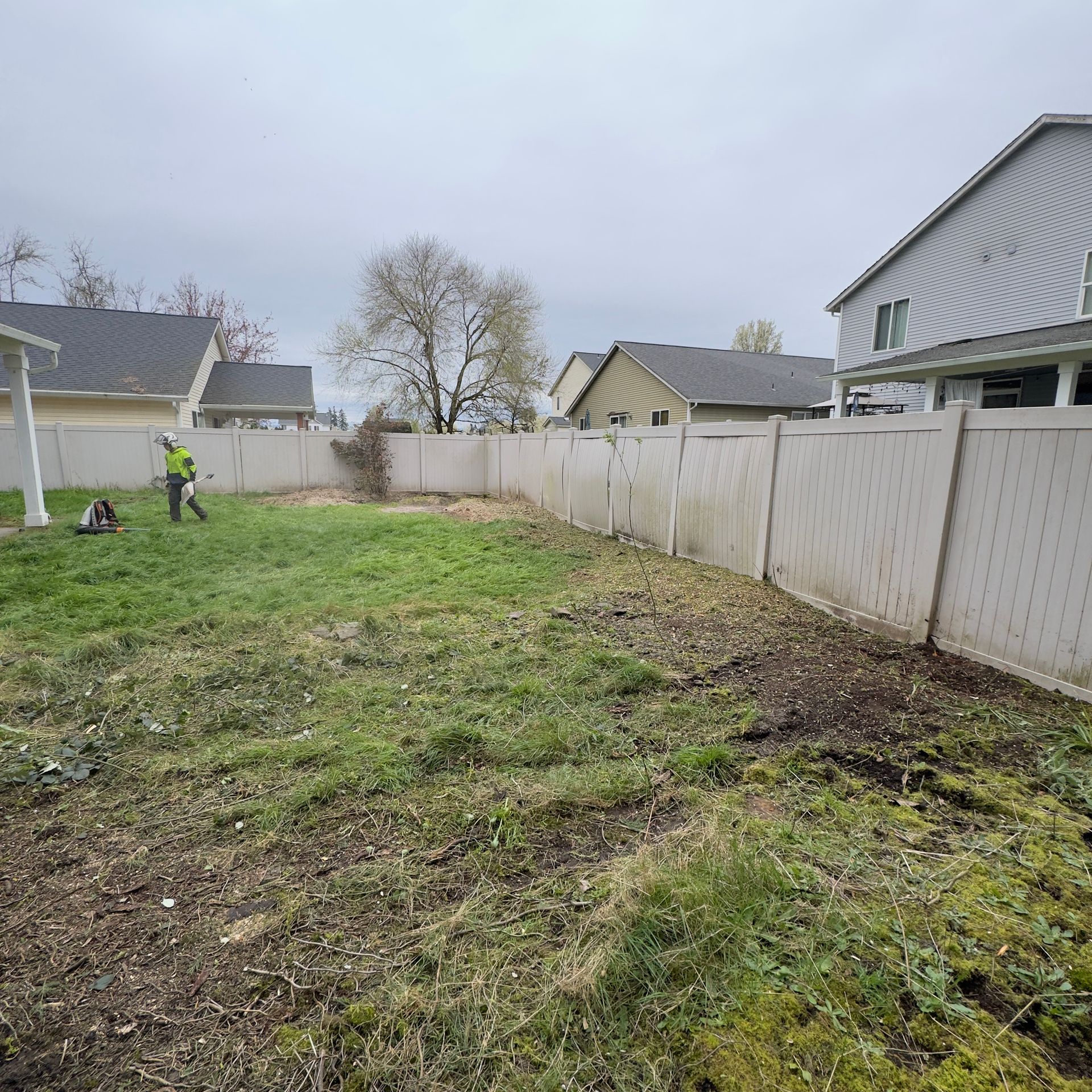 Backyard with muddy grass, a worker in a neon jacket, and a white fence under a cloudy sky