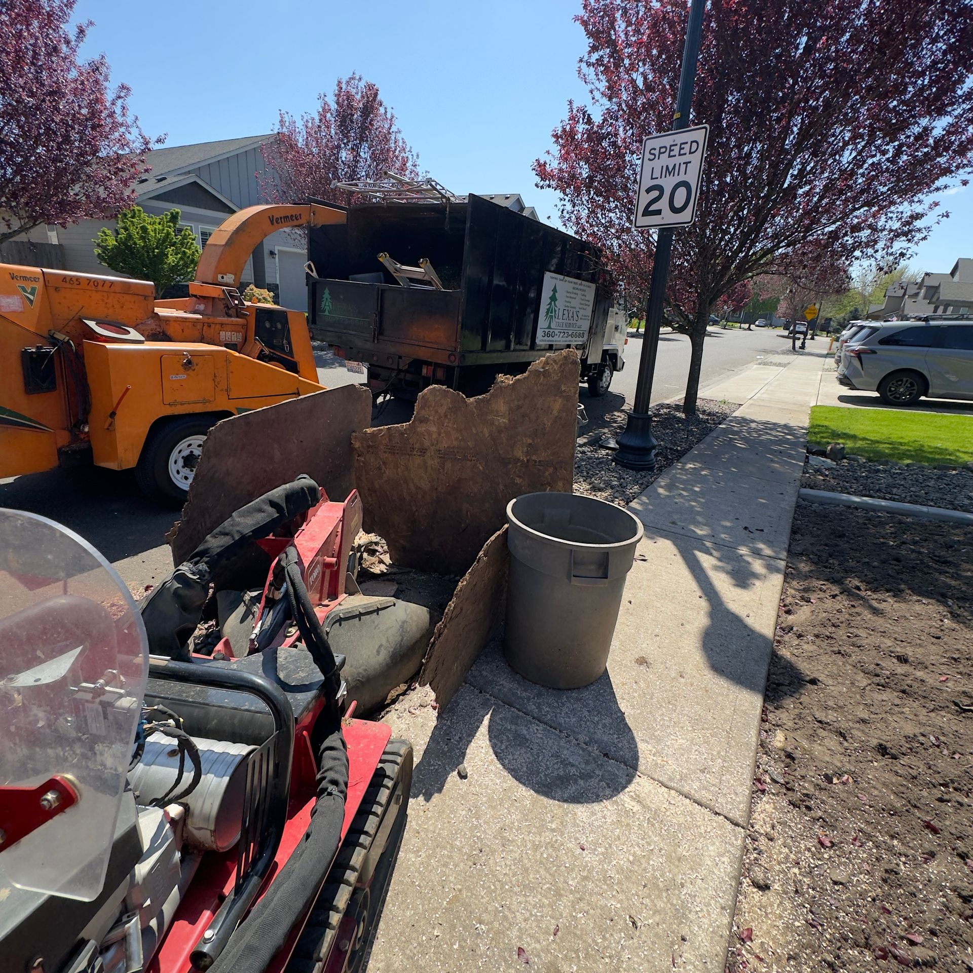 Construction site with orange machinery, dumpster, and trash bins on a sidewalk beside trees and parked cars