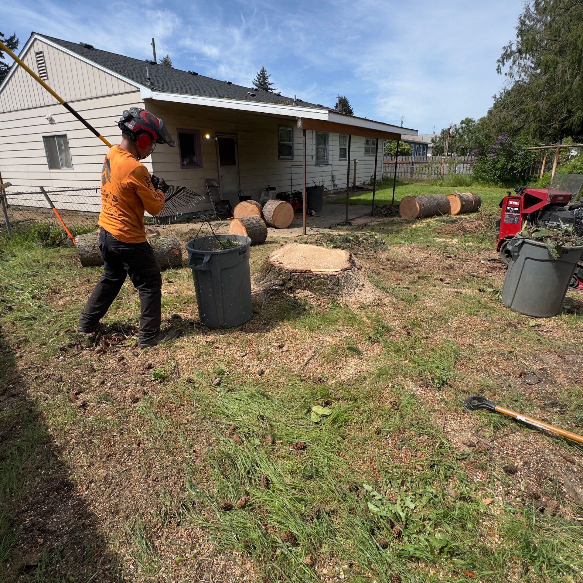 Worker using a chainsaw beside a house, with logs, a trash bin, and cut branches in the yard.