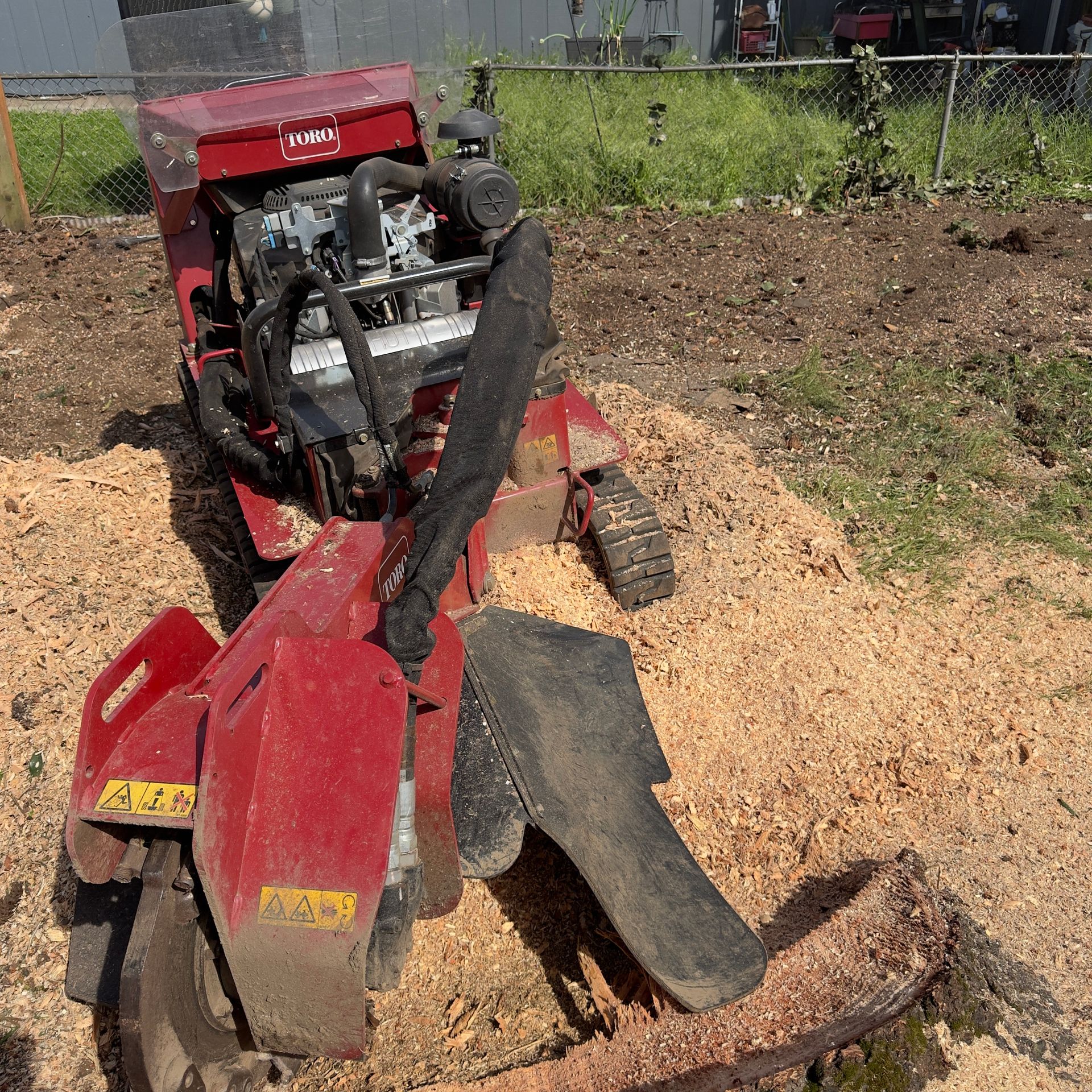 Red farm tiller with attached plow sitting in a dirt field beside a garden fence