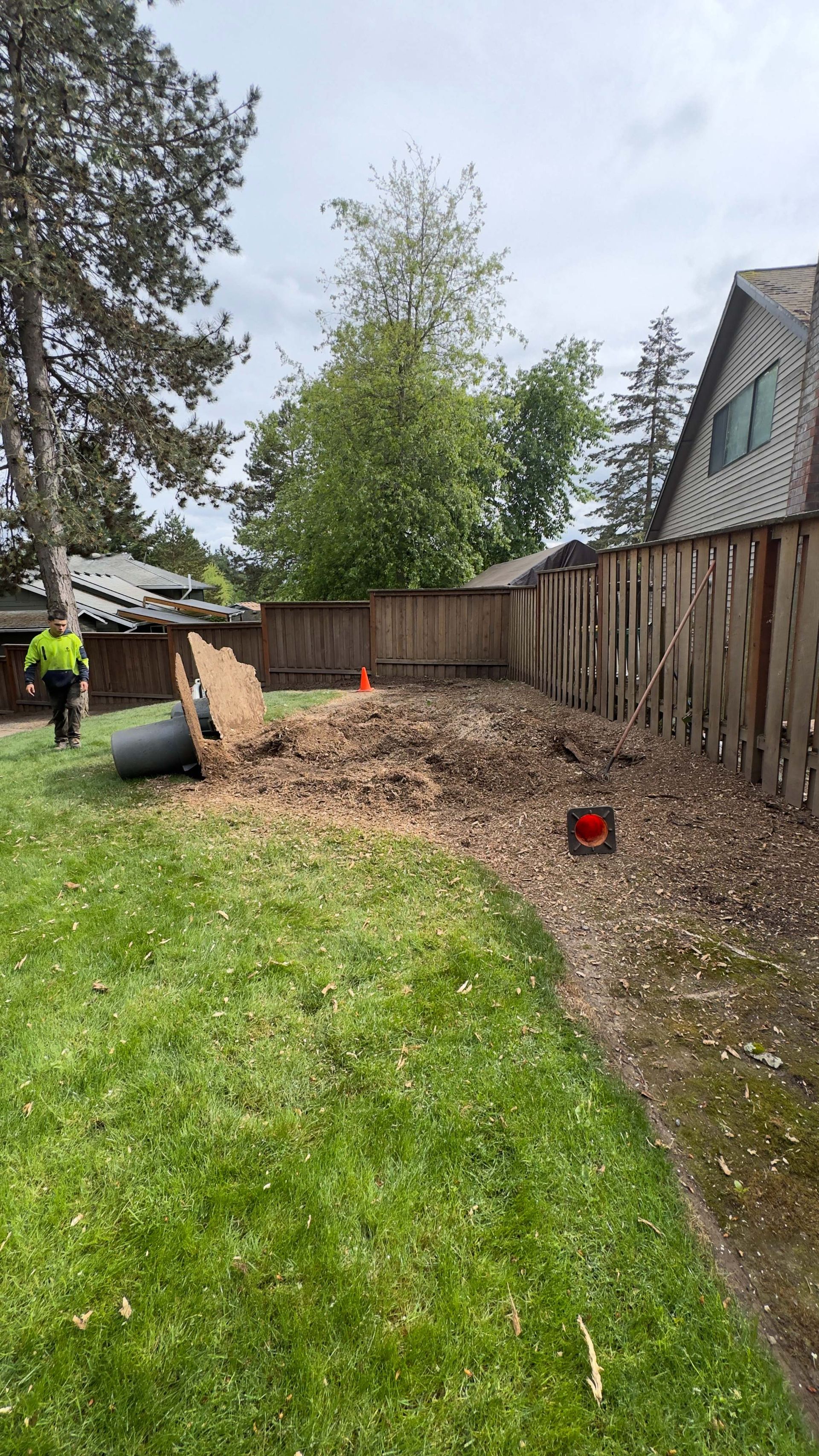 Person in high-visibility jacket works in a grassy backyard beside a fence and wood chips, with tools nearby