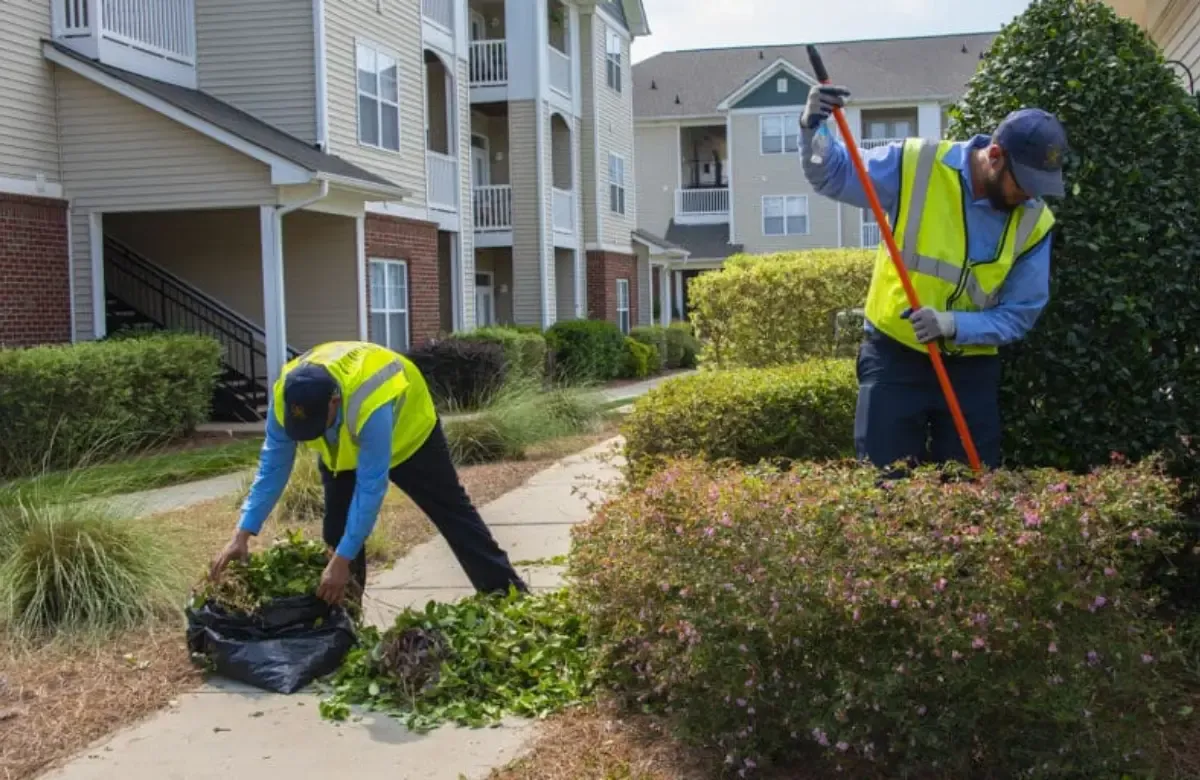 Two workers in yellow safety vests clear leaves along a sidewalk in an apartment complex