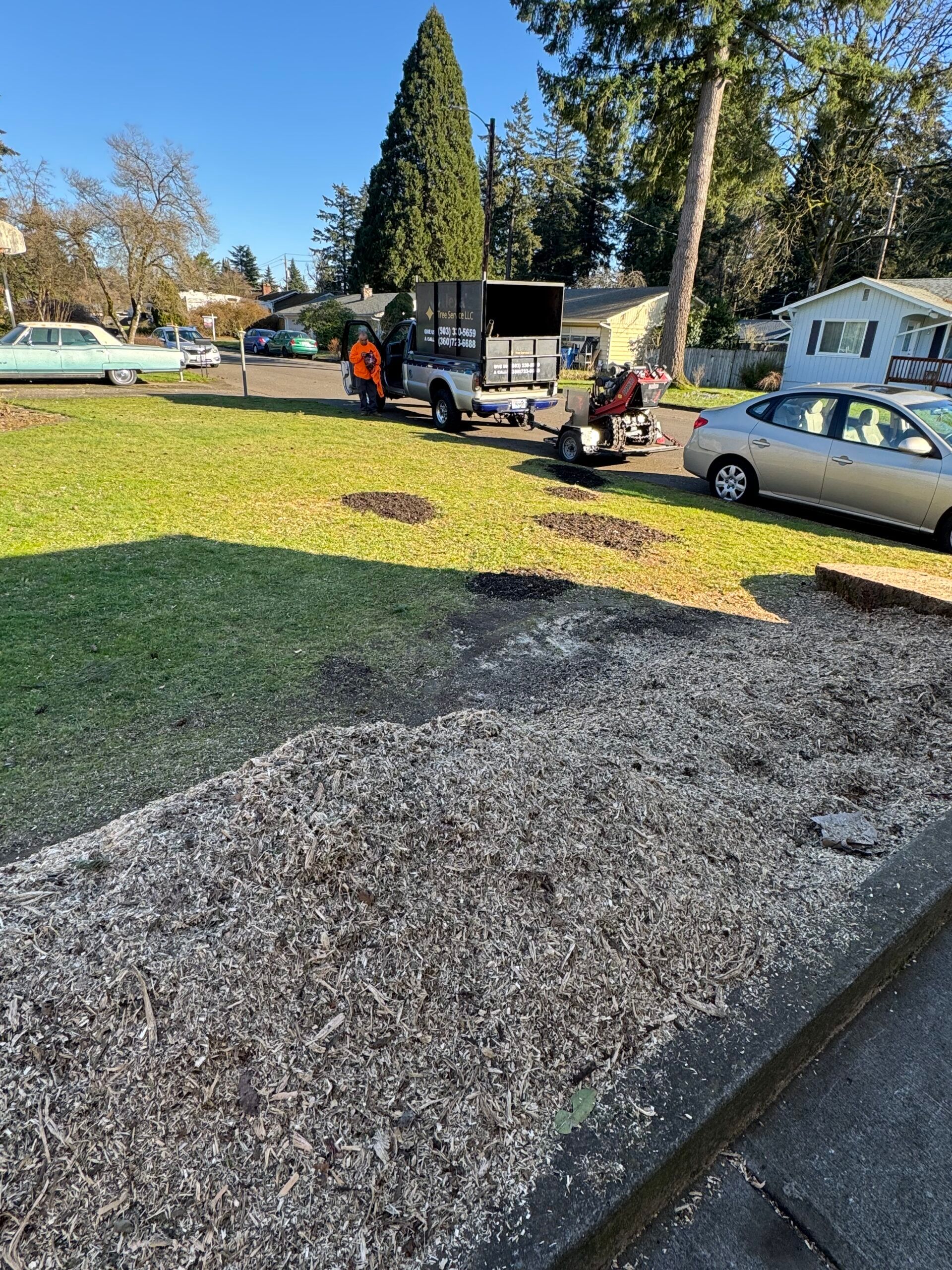 Gravel driveway beside a green lawn with workers, a utility truck, and parked cars on a sunny day