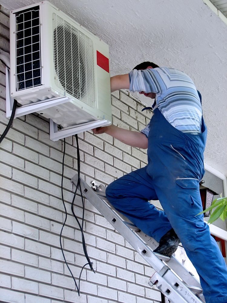 A man is standing on a ladder fixing an air conditioner on a brick wall.