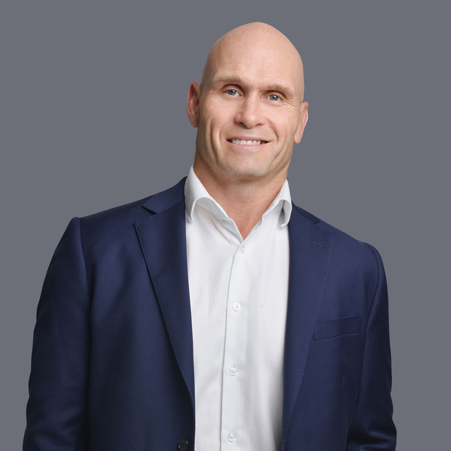 Bald man wearing a navy blazer and white shirt, smiling against a gray background.