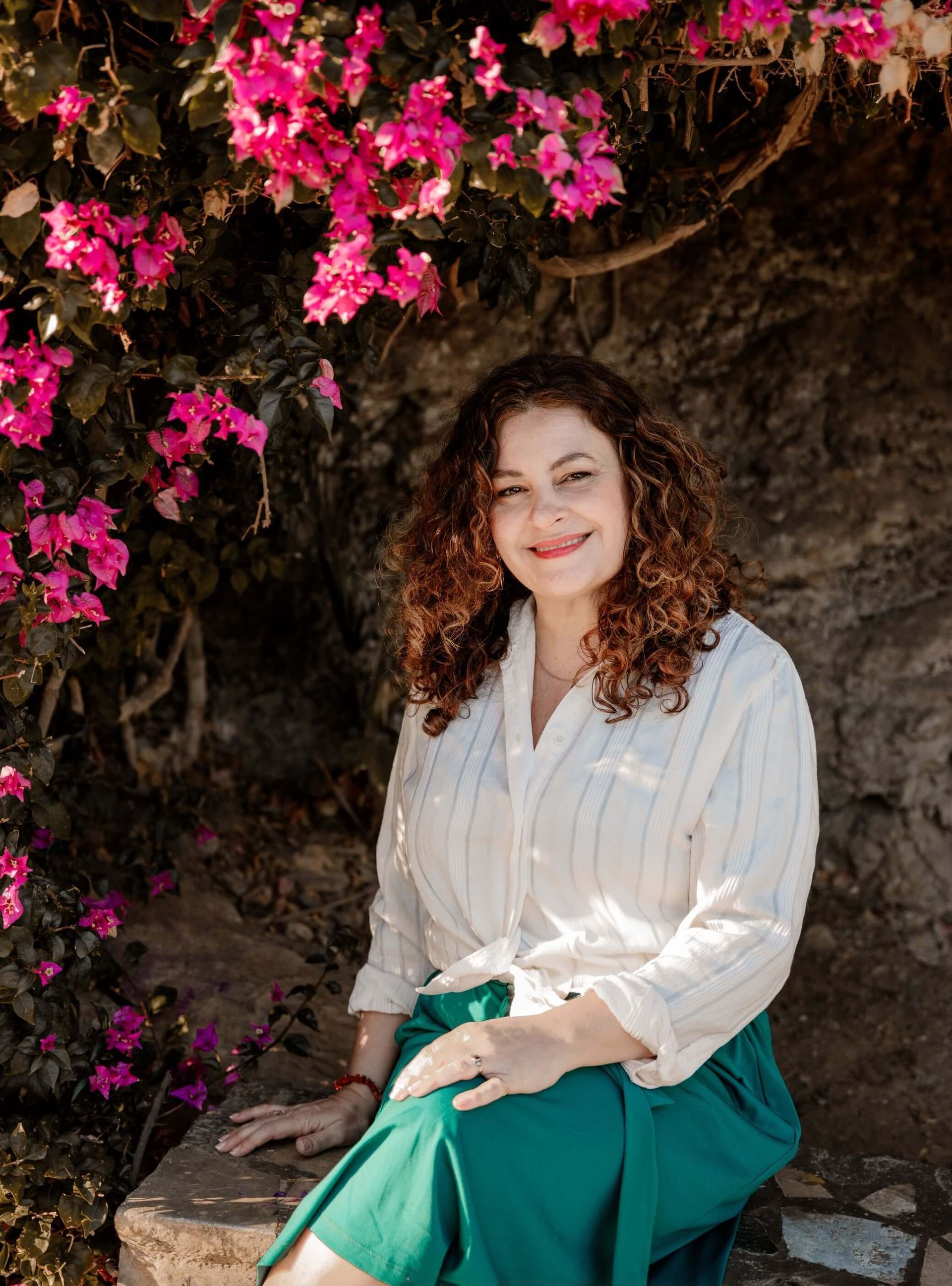 Mónica García Hidalgo está sentada en una roca frente a un arbusto con flores rosas.