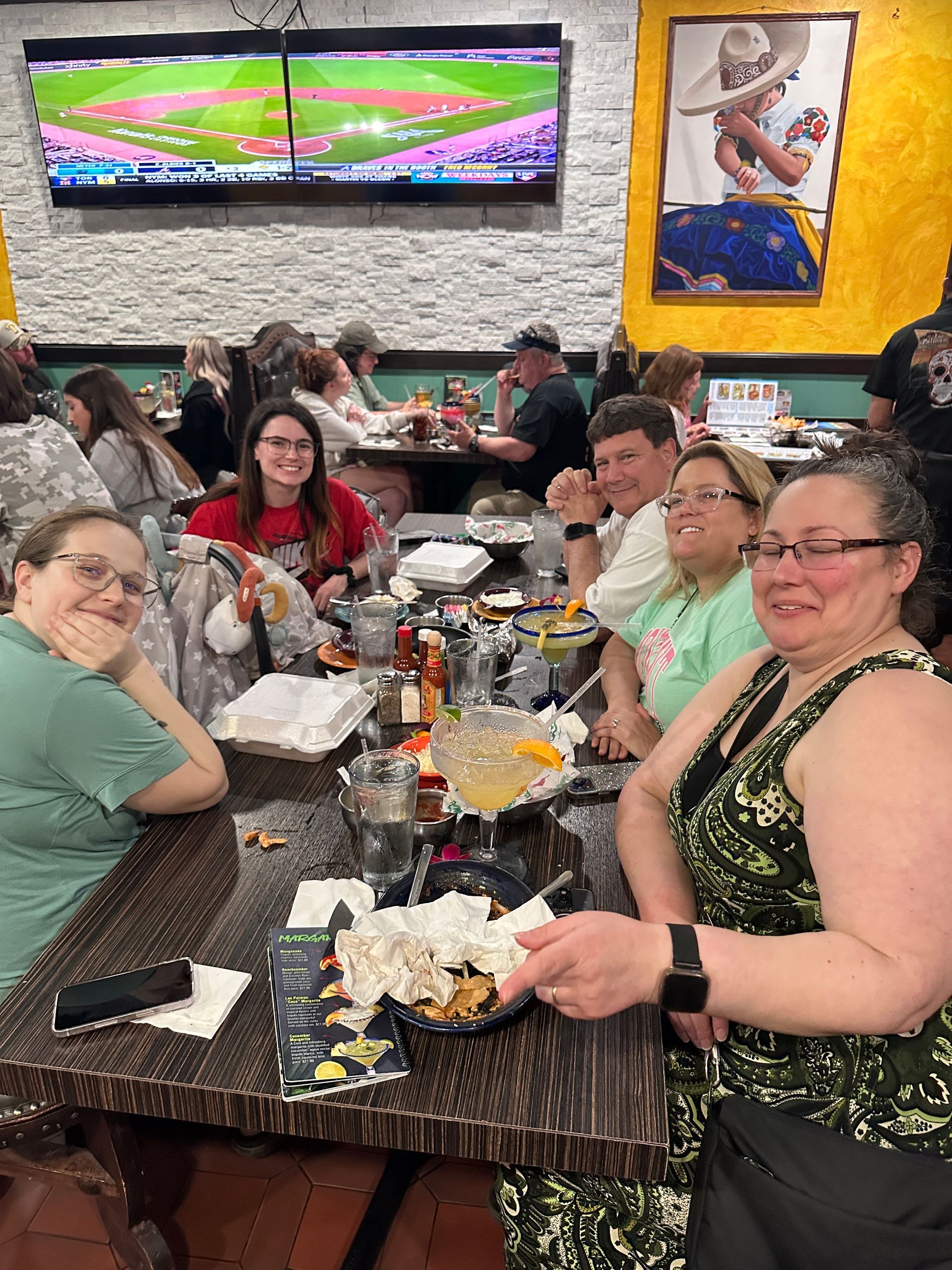 People at a restaurant table, smiling, with food and drinks. Baseball game on TV in background.