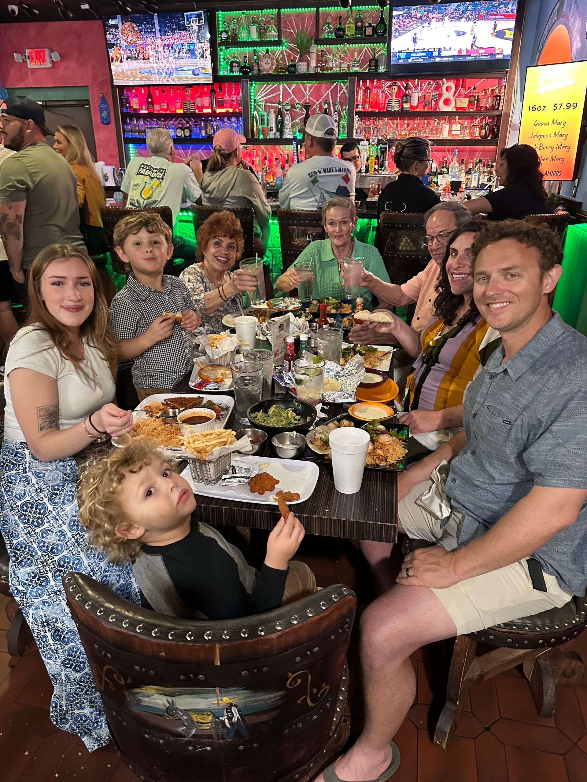 Group of people at a restaurant table with food; colorful bar and TVs in background.