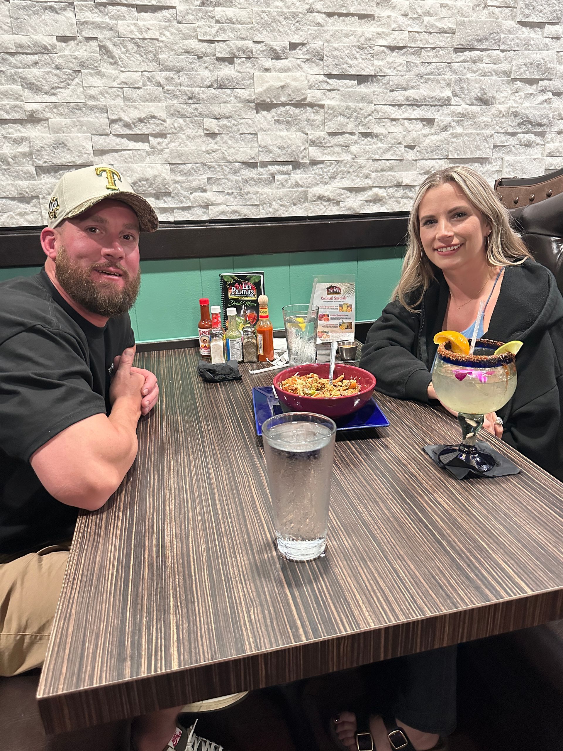 Two people at a restaurant table. Man in hat and woman with cocktail. Food and drinks are present.