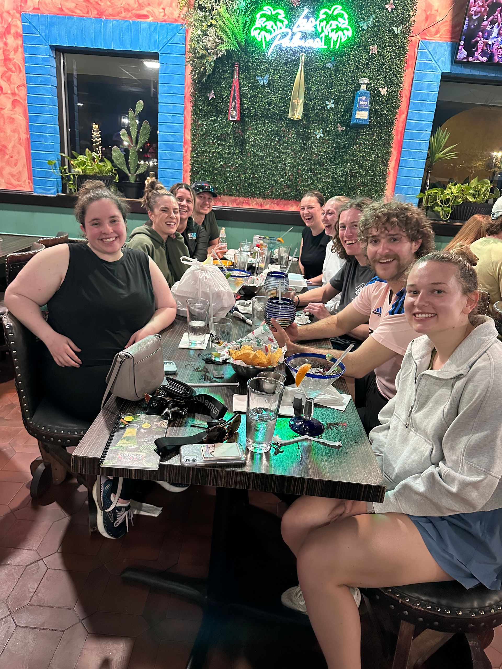 Group of people seated at a table in a Mexican restaurant, smiling. Chips, salsa, and drinks are visible.