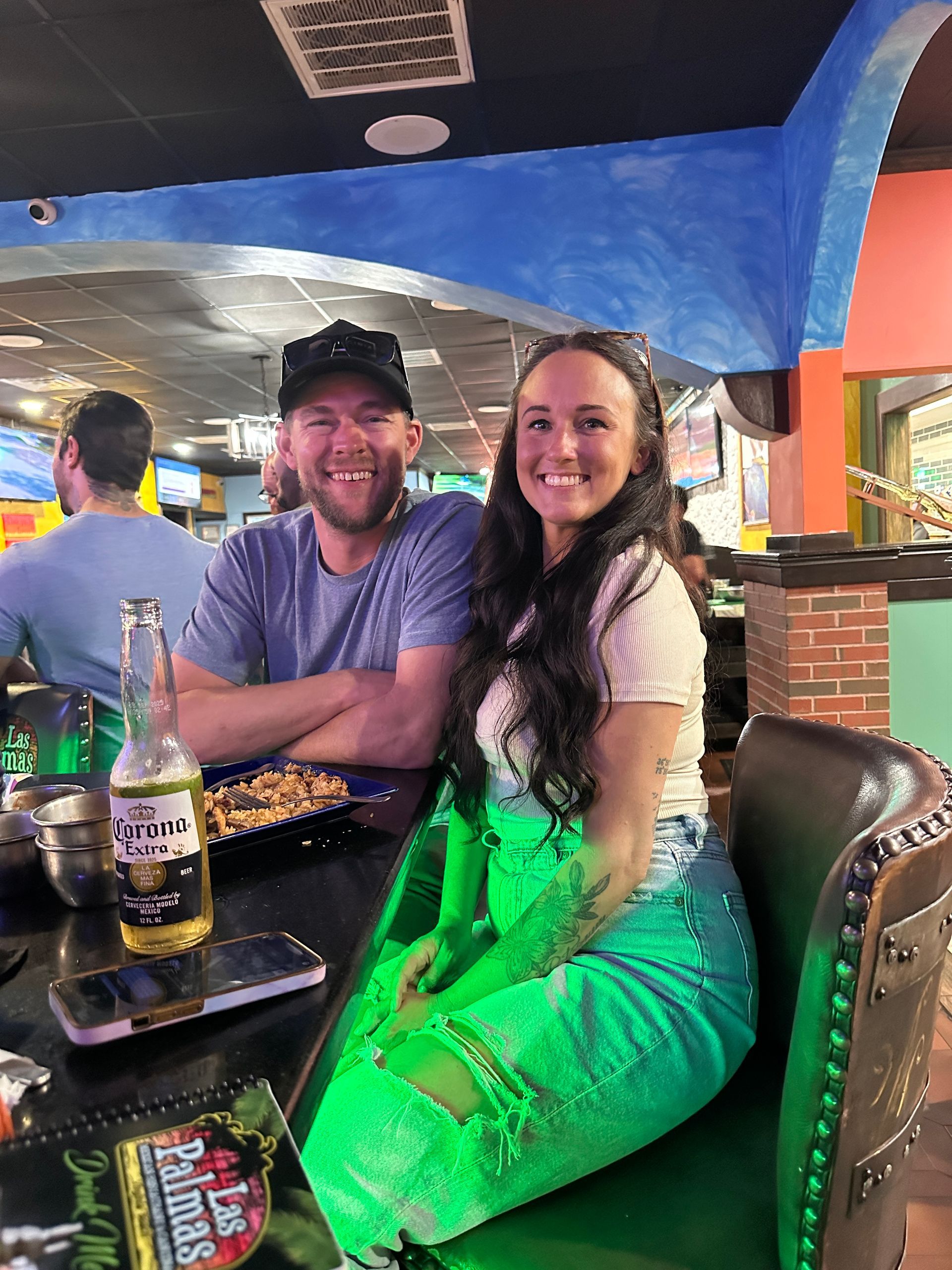 Couple smiling at a restaurant, seated at a table with beer and a phone. Green and blue decor.