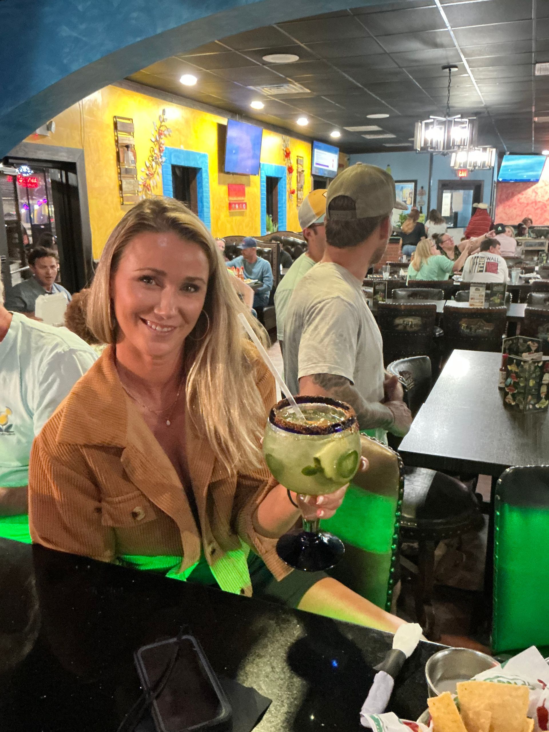 Woman at a bar holding a margarita. She smiles, wearing a tan jacket, and has long blonde hair. Interior shot of a Mexican restaurant.