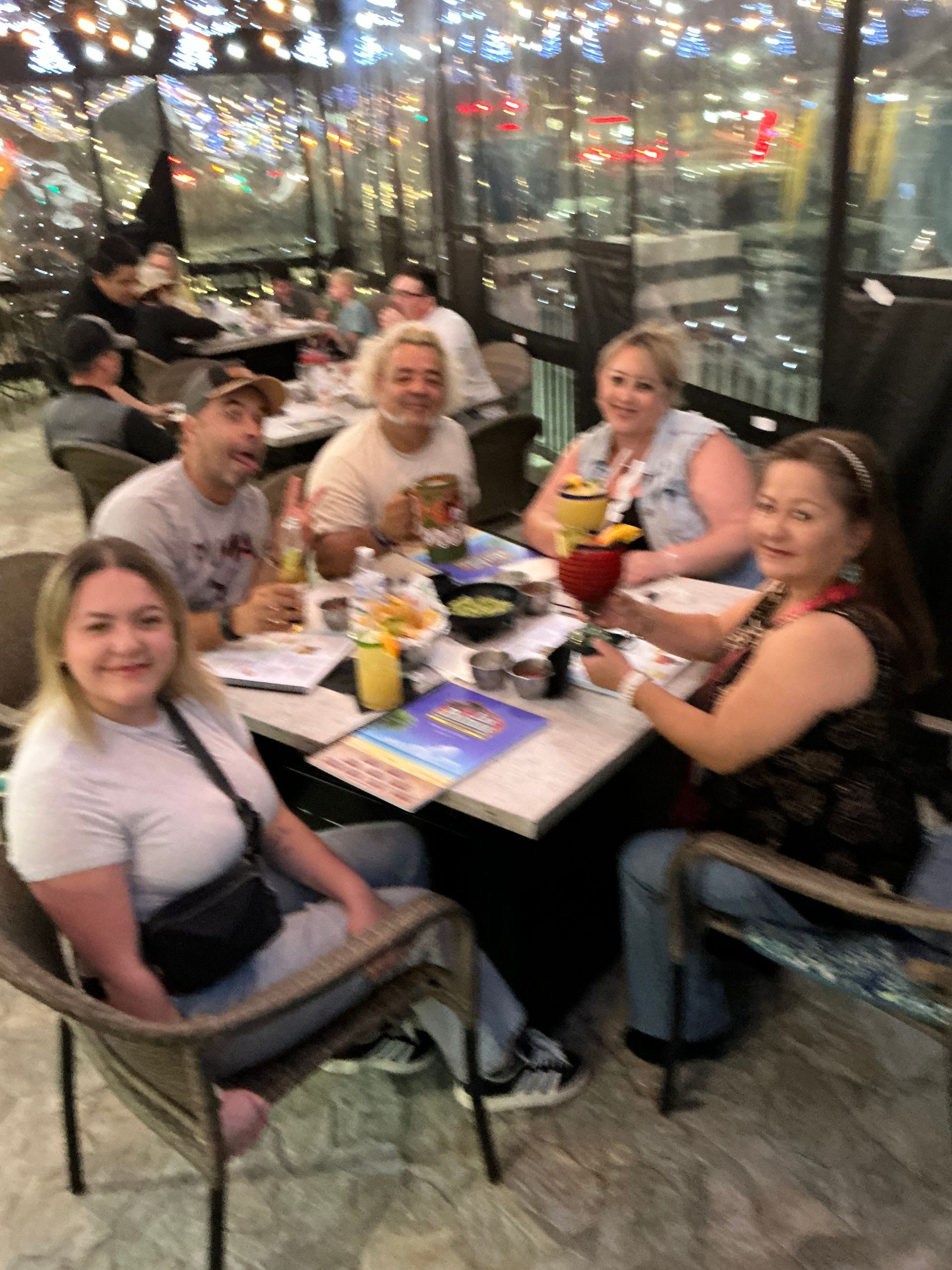 Group of people seated at a table, some holding drinks, in an outdoor restaurant setting.