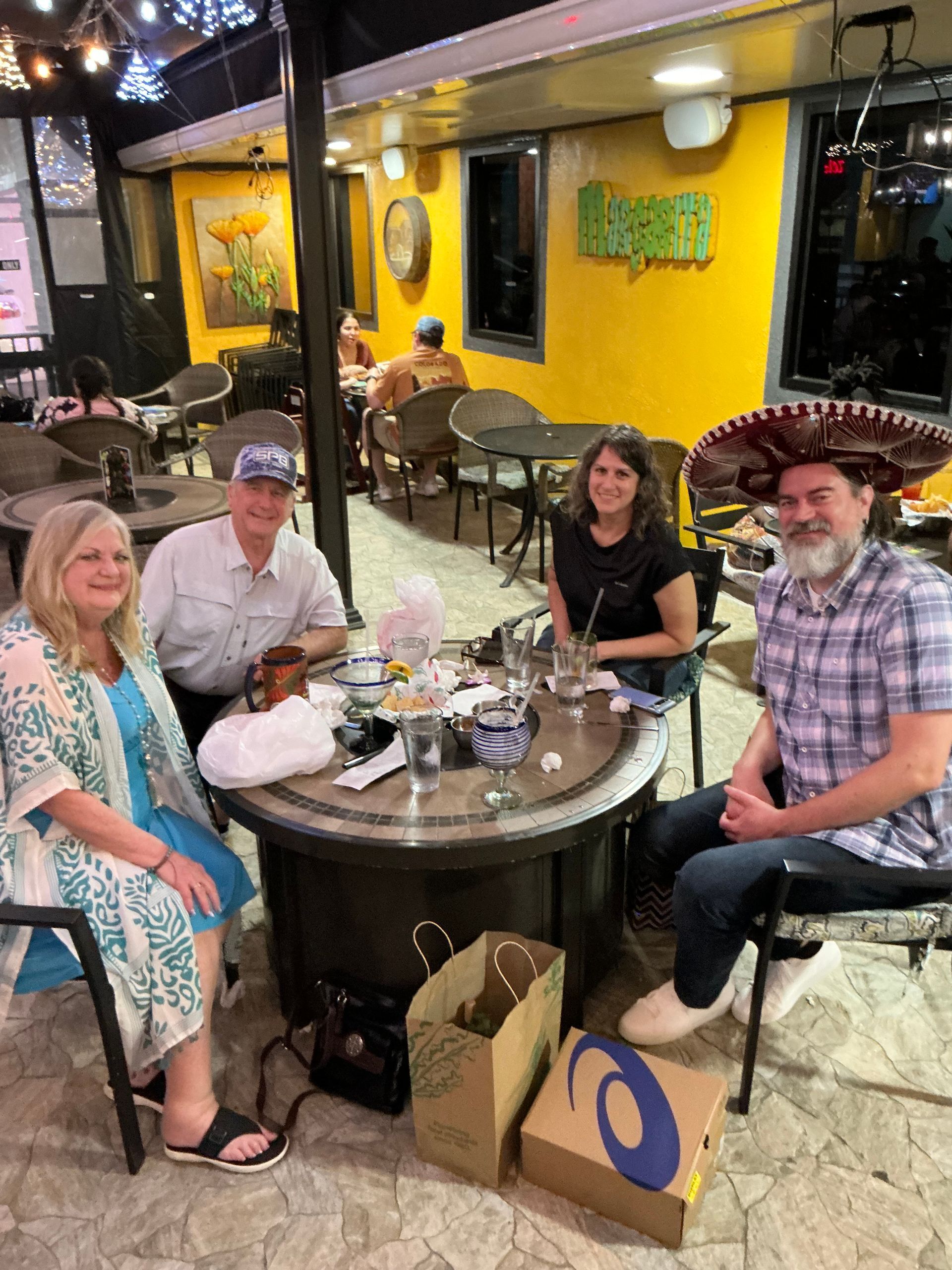 Four people seated around a round table at a restaurant, one wearing a sombrero. Yellow walls and outdoor setting.